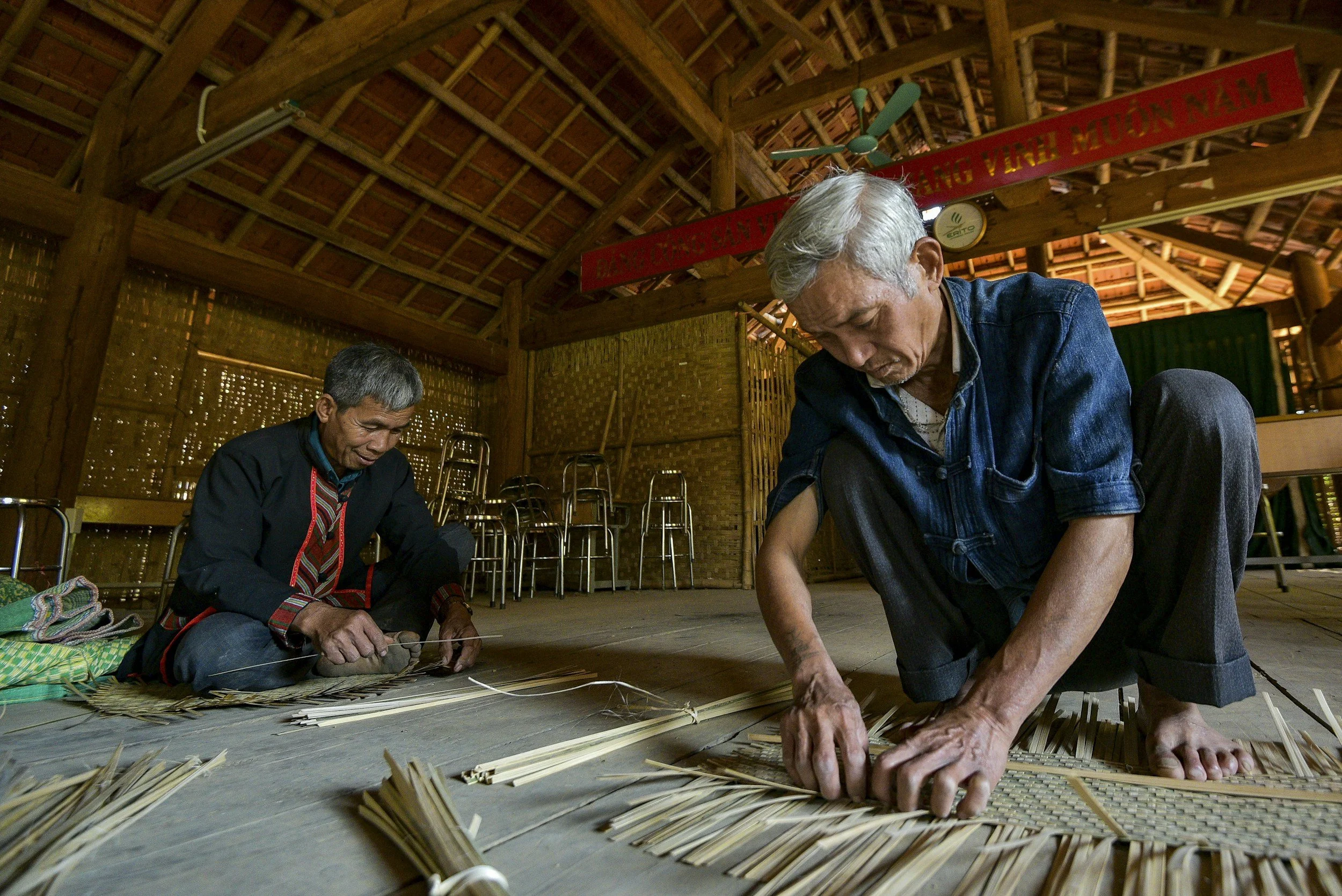Two men working on weaving mats with straw inside a wooden building.