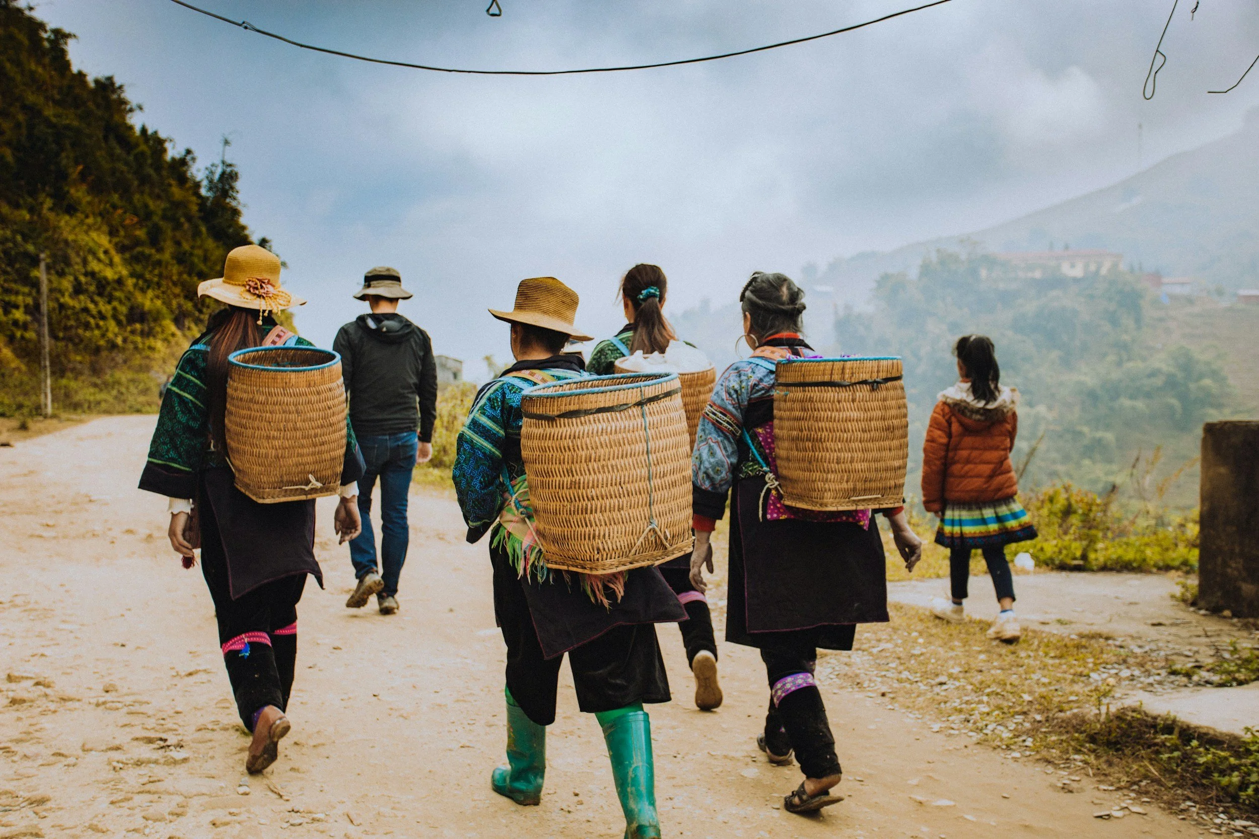 Group of people walking along a dirt path in a rural, mountainous area, some wearing traditional clothing and carrying large woven baskets on their backs.