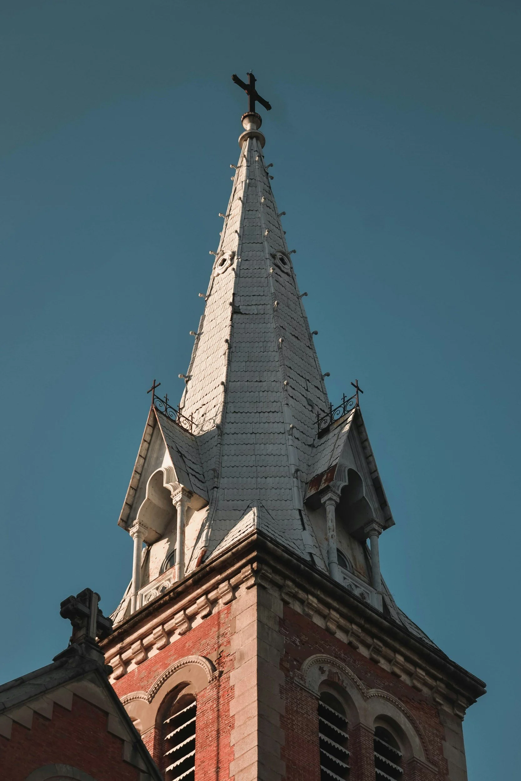 The steeple of a church building with a cross at the top, featuring pointed roofing and decorative architectural details, against a clear blue sky.