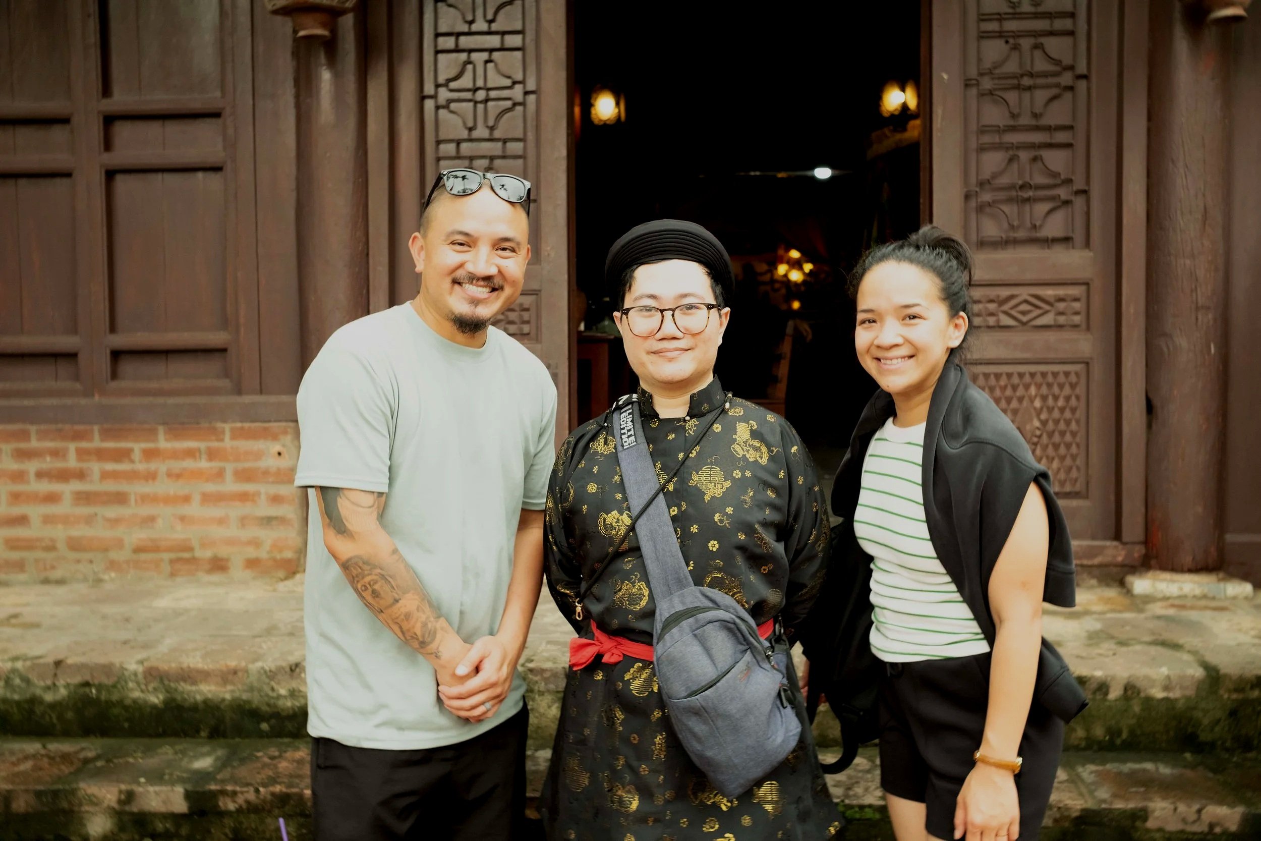 Three people smiling outdoors in front of a wooden door with Asian architectural details. The man on the left has glasses on his head, a beard, and tattoos on his arm. The person in the middle has glasses, a hat, and a camera strap around their neck.