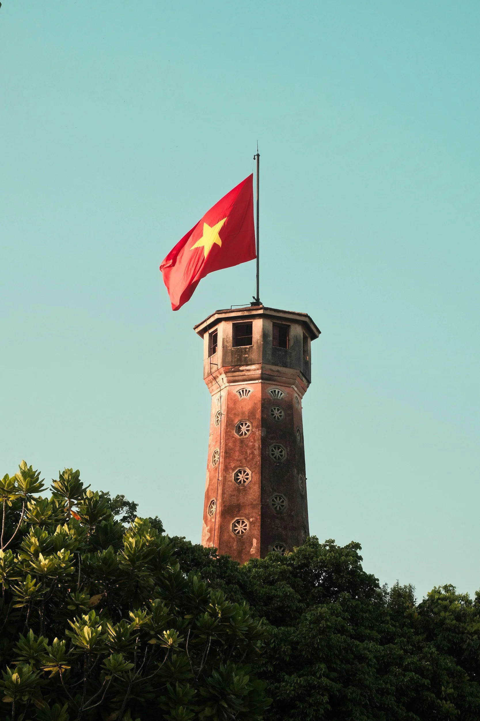 A tall historical tower with a flagpole at the top flying a red flag with a yellow star, surrounded by lush green trees, against a clear sky.