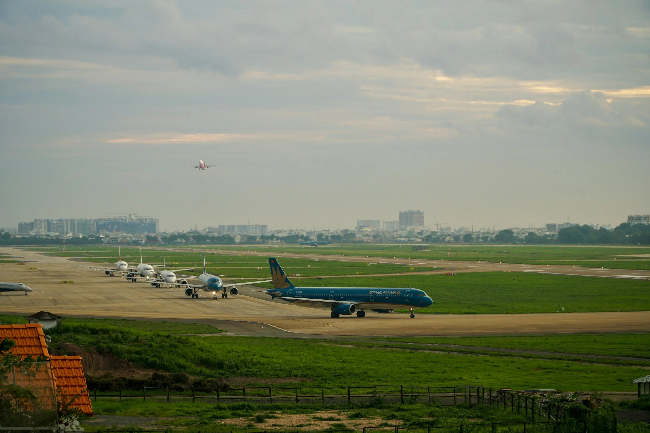 View of an airport with several airplanes parked on the tarmac, including a Vietnam Airlines plane, and an airplane taking off in the sky under a cloudy sky with city buildings in the background.