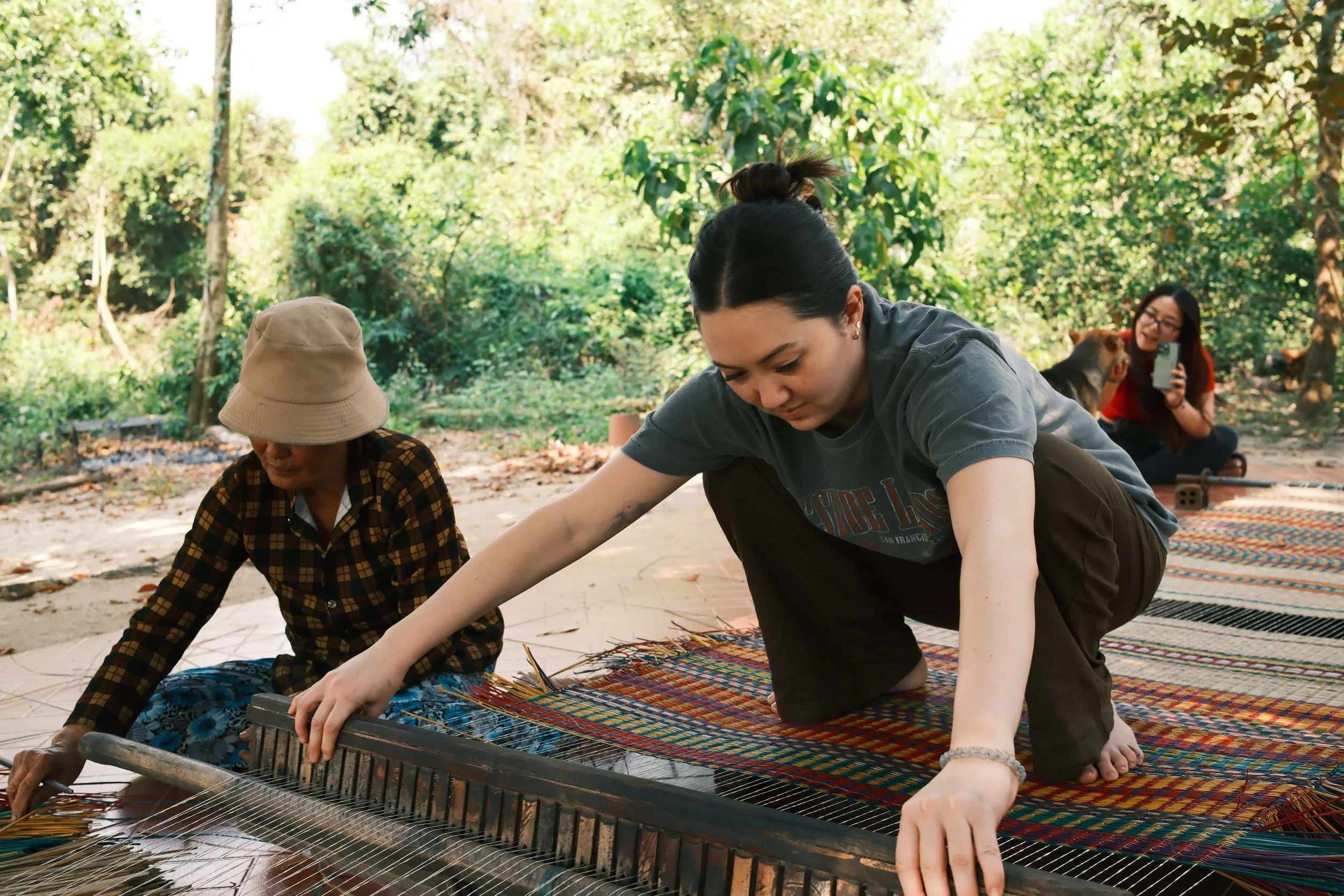 Group of people engaging in a traditional craft or art project outdoors in a lush, green setting. They are working on a colorful woven textile laid out on the ground, with some bending over and focusing on their tasks.