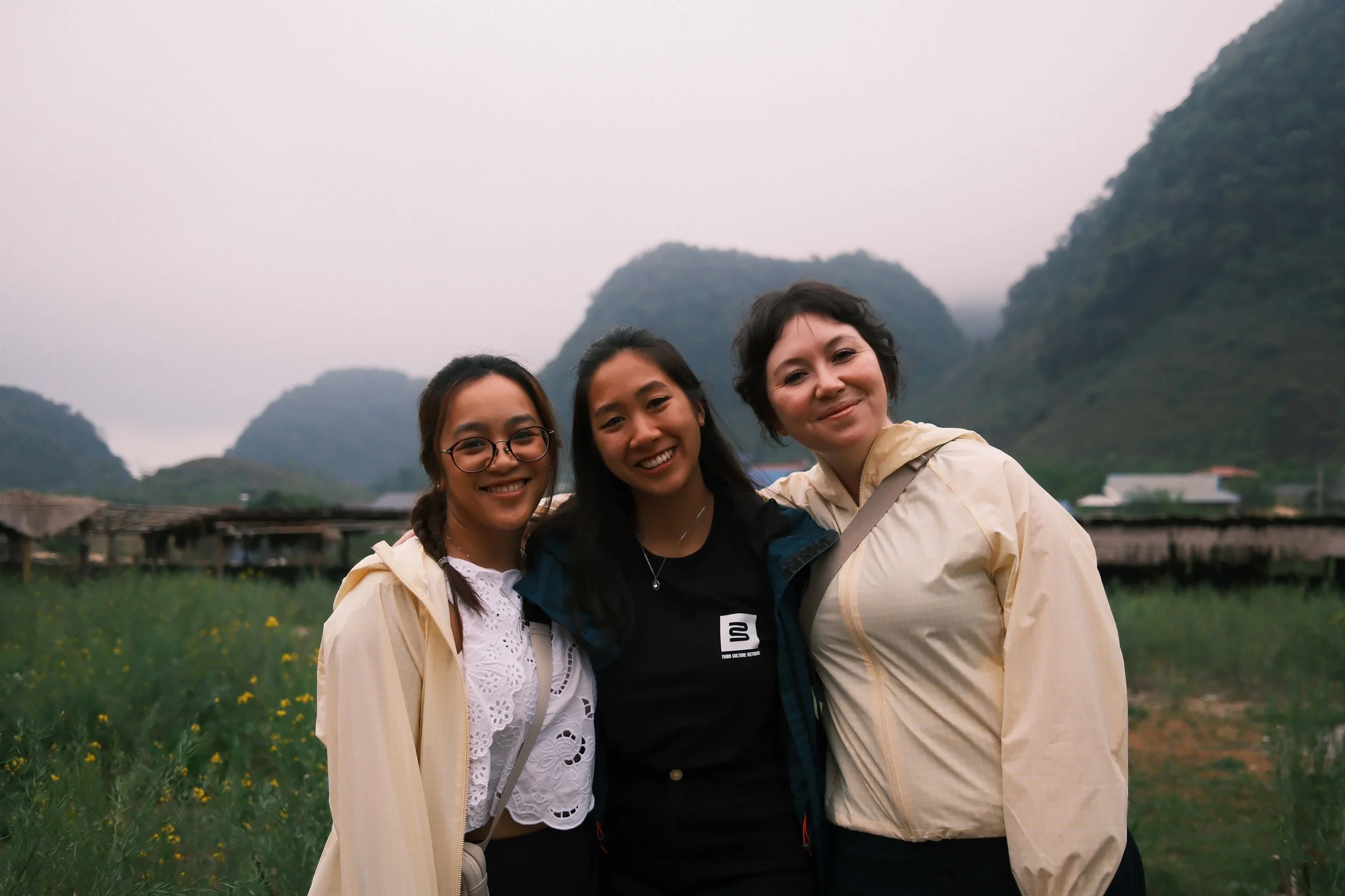 Three women standing outdoors in a field with hills in the background, smiling at the camera.