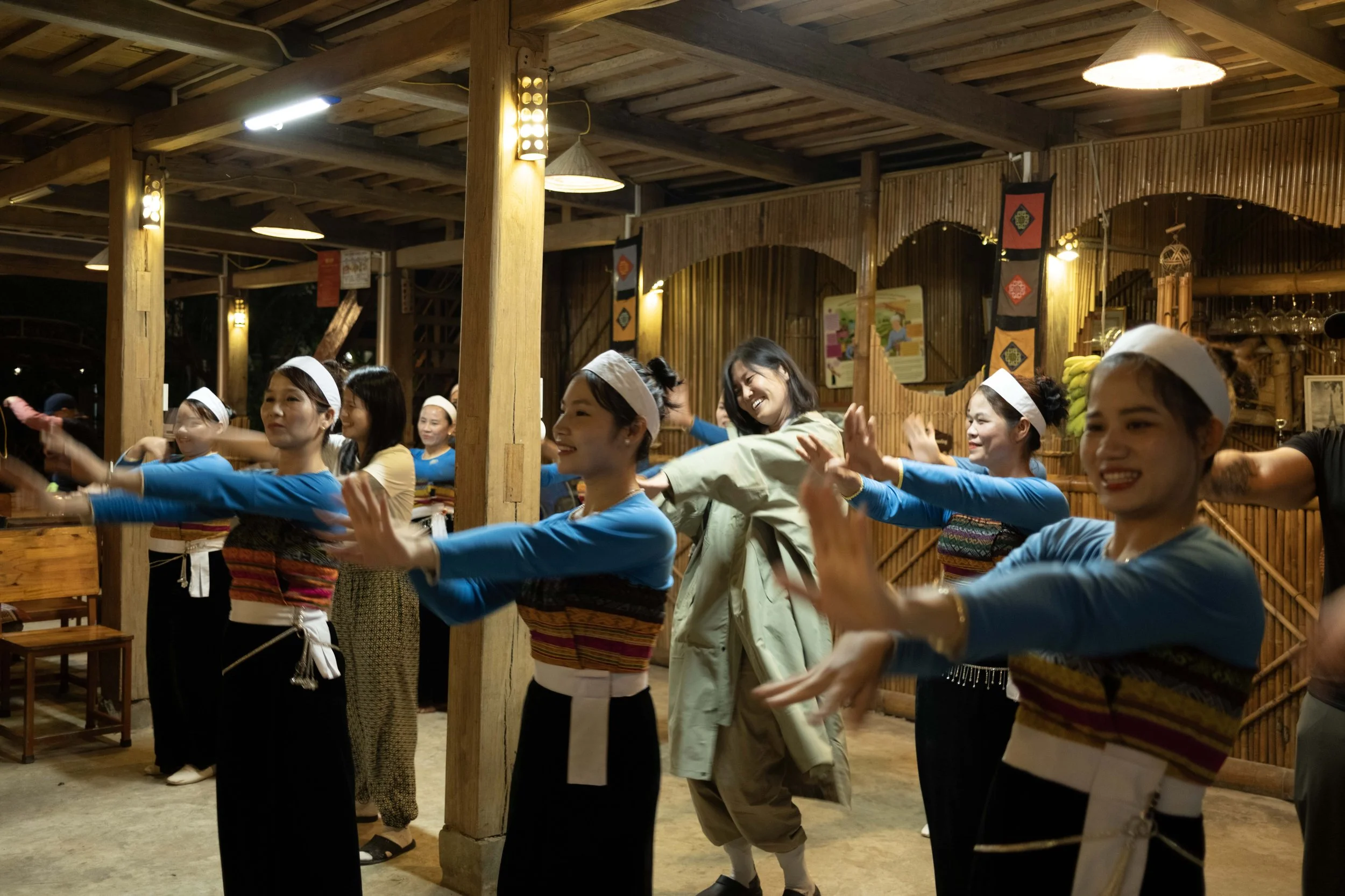 Group of women dancing in a traditional or cultural setting, wearing matching blue tops, white headbands, and patterned skirts, inside a wooden venue with warm lighting.
