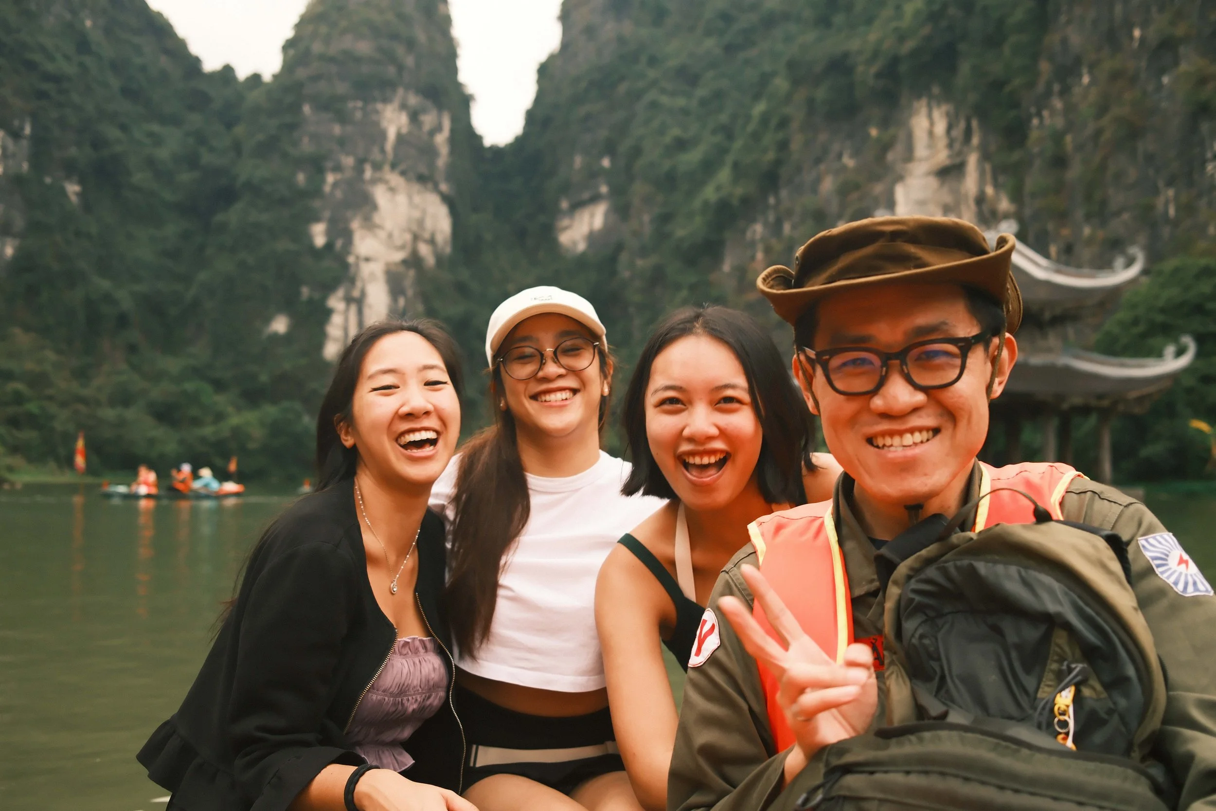 A group of four smiling diverse friends taking a selfie outdoors near a river surrounded by lush green mountains, with a boat visible in the background.