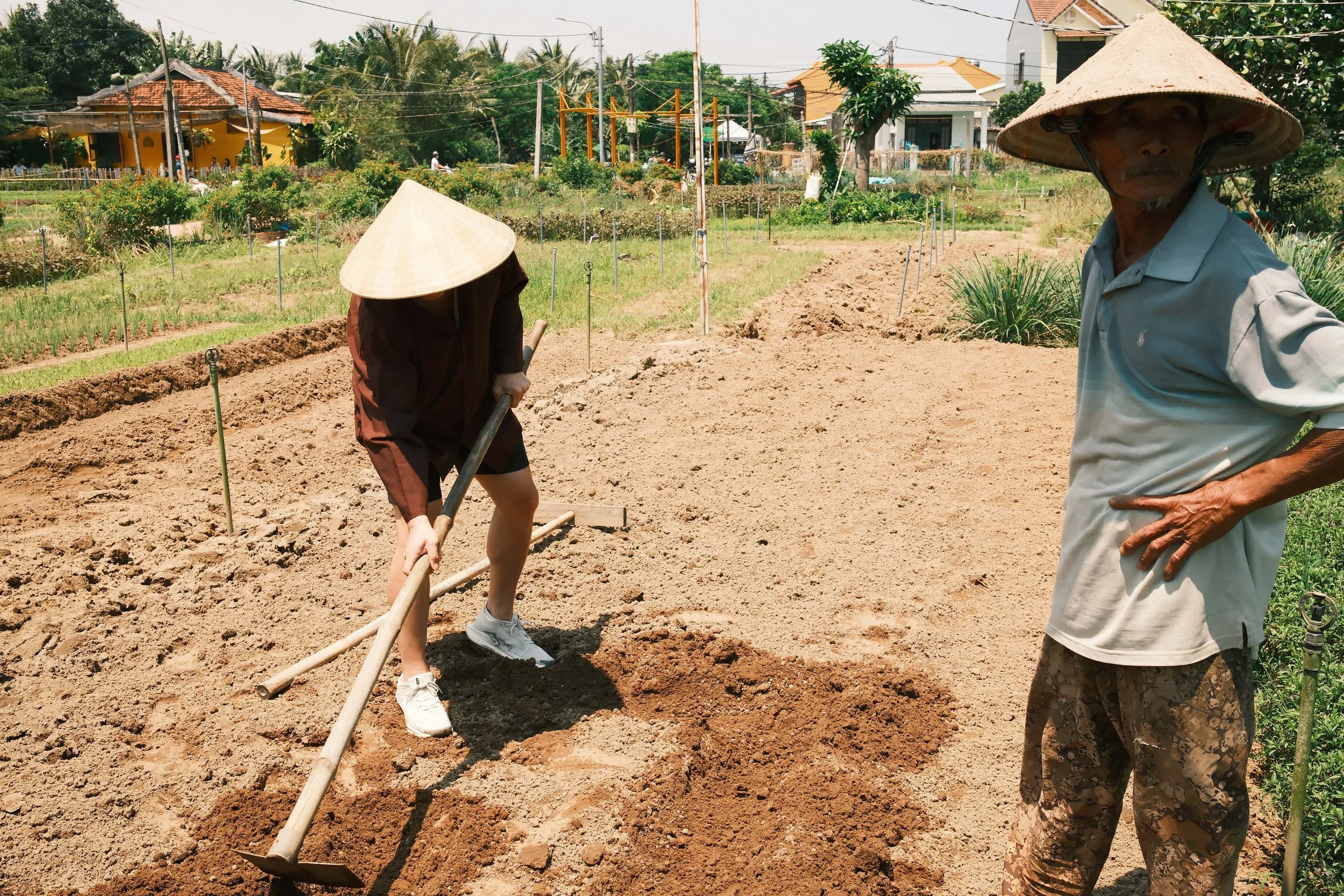 Two individuals, one with a conical hat and casual clothing, working in a garden or farm, using a tool to till or prepare the soil; another person, also with a conical hat, standing nearby with hands on hips, overlooking the activity, in a rural area