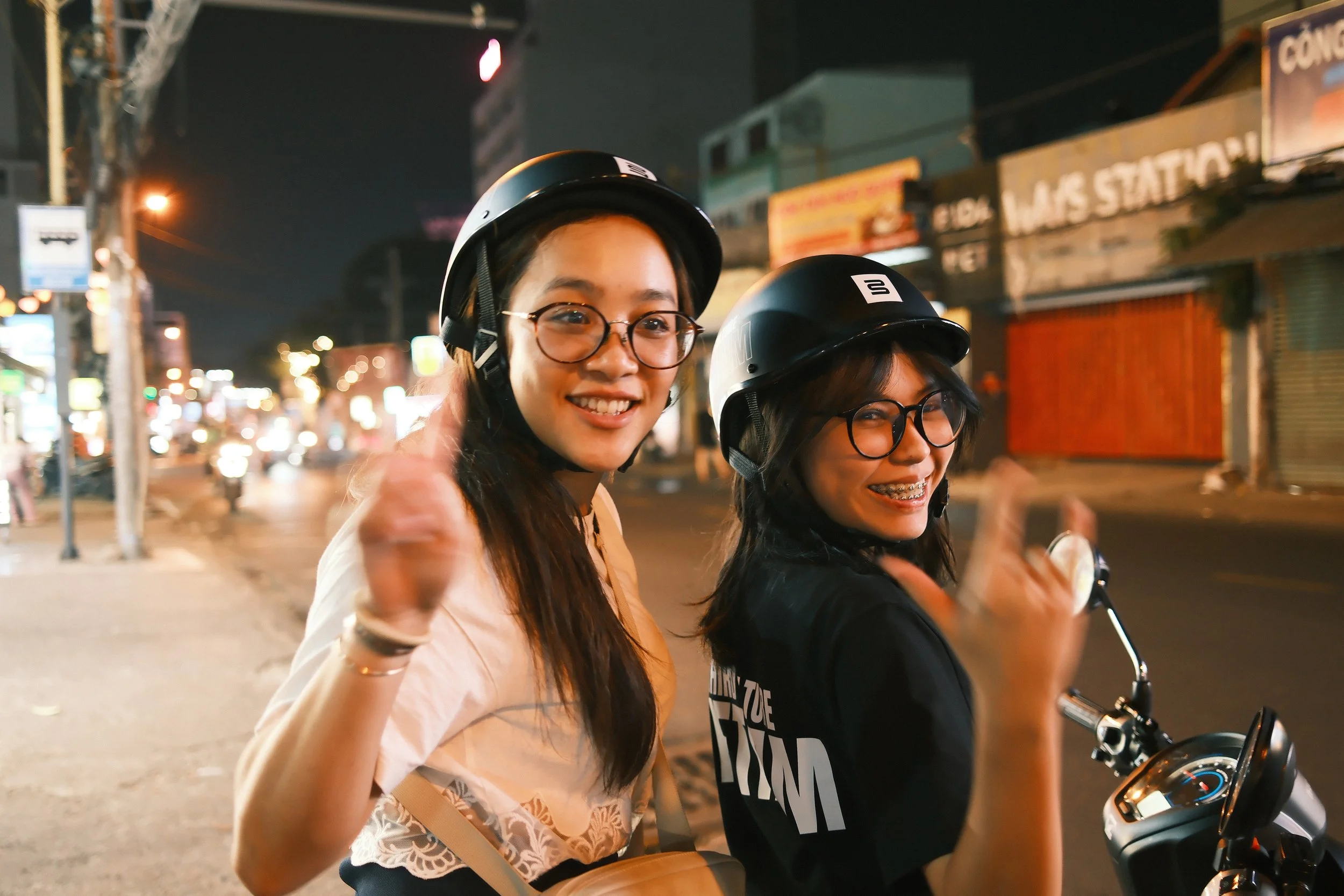 Two young women wearing helmets on a scooter at night in an urban setting, smiling and waving.