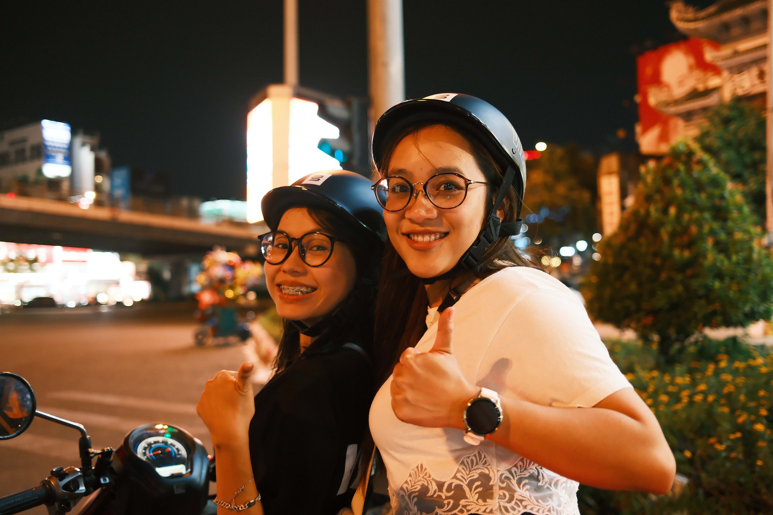 Two women wearing helmets sitting on a motorcycle at night in a city setting, smiling and giving thumbs-up gestures.