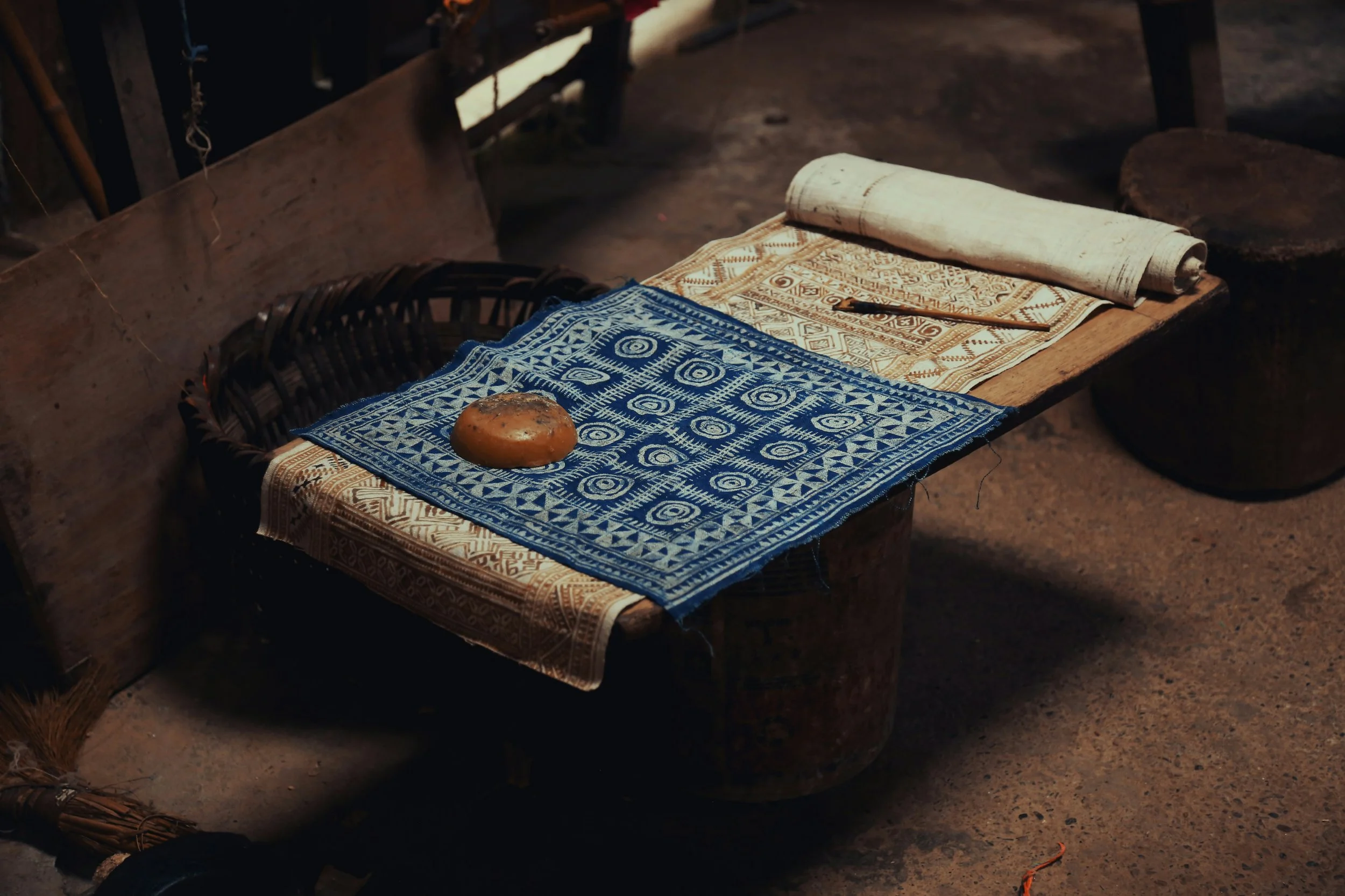 Traditional Vietnamese woven textiles on a rustic wooden table with a stone in the center, and rolled fabrics in the background.