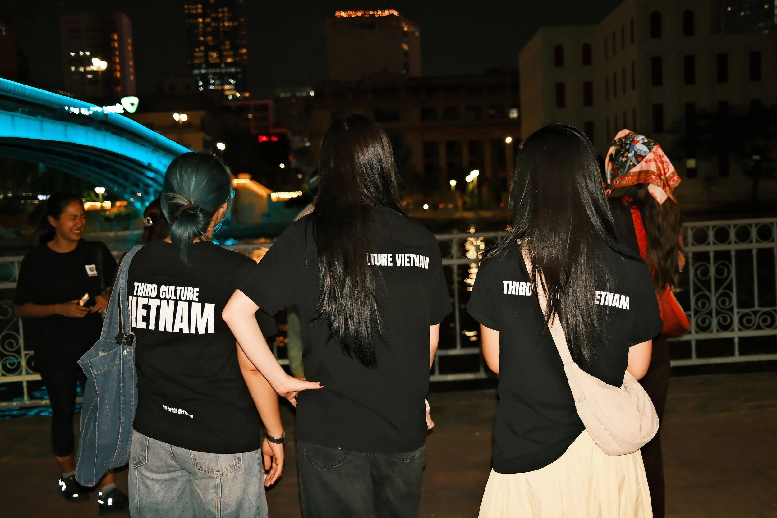 Group of young women wearing matching black T-shirts with white text that says 'THIRD CULTURE VIETNAM' on a nighttime cityscape by the water, with illuminated buildings and a bridge in the background.