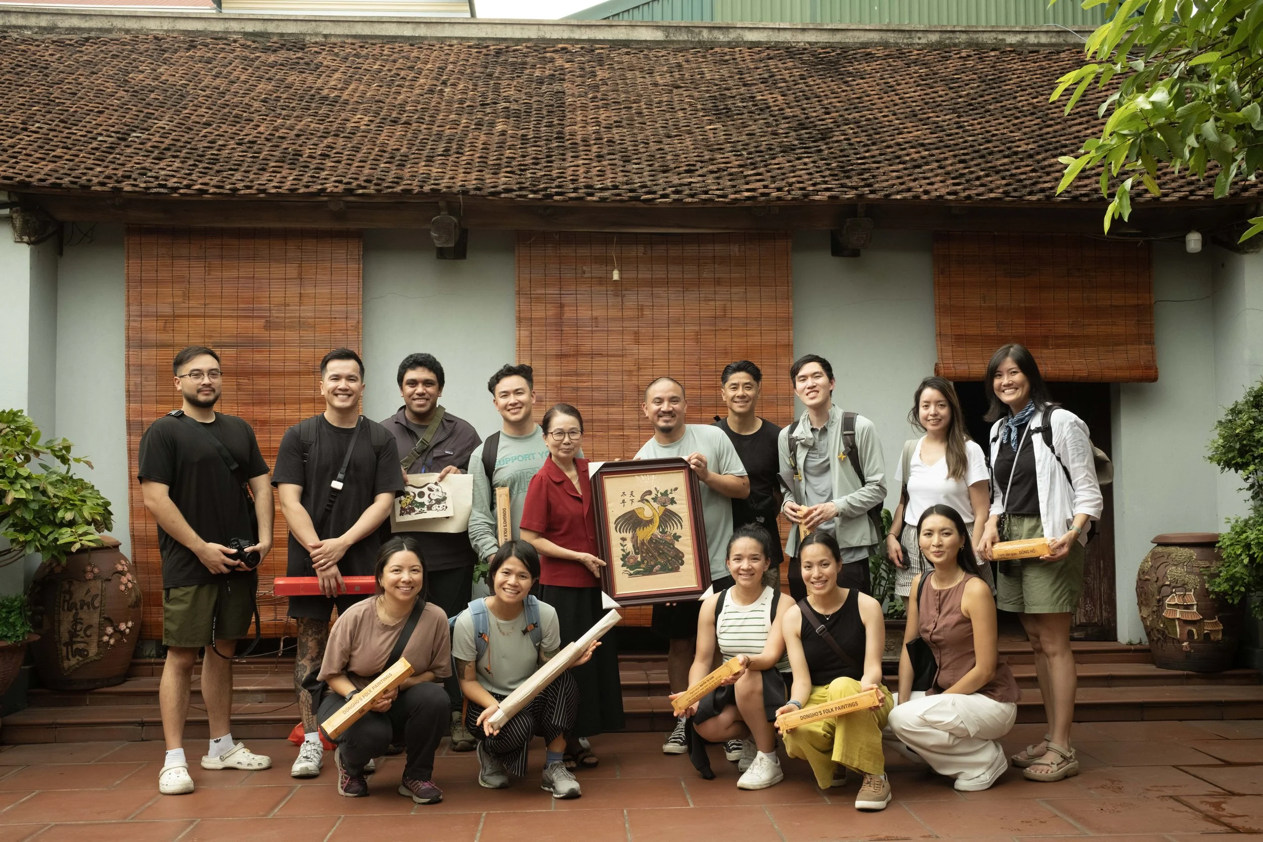 Group of people posing on a wooden deck in front of a building with bamboo blinds and greenery. Some individuals hold scrolls, and one person holds a framed artwork with a peacock design.