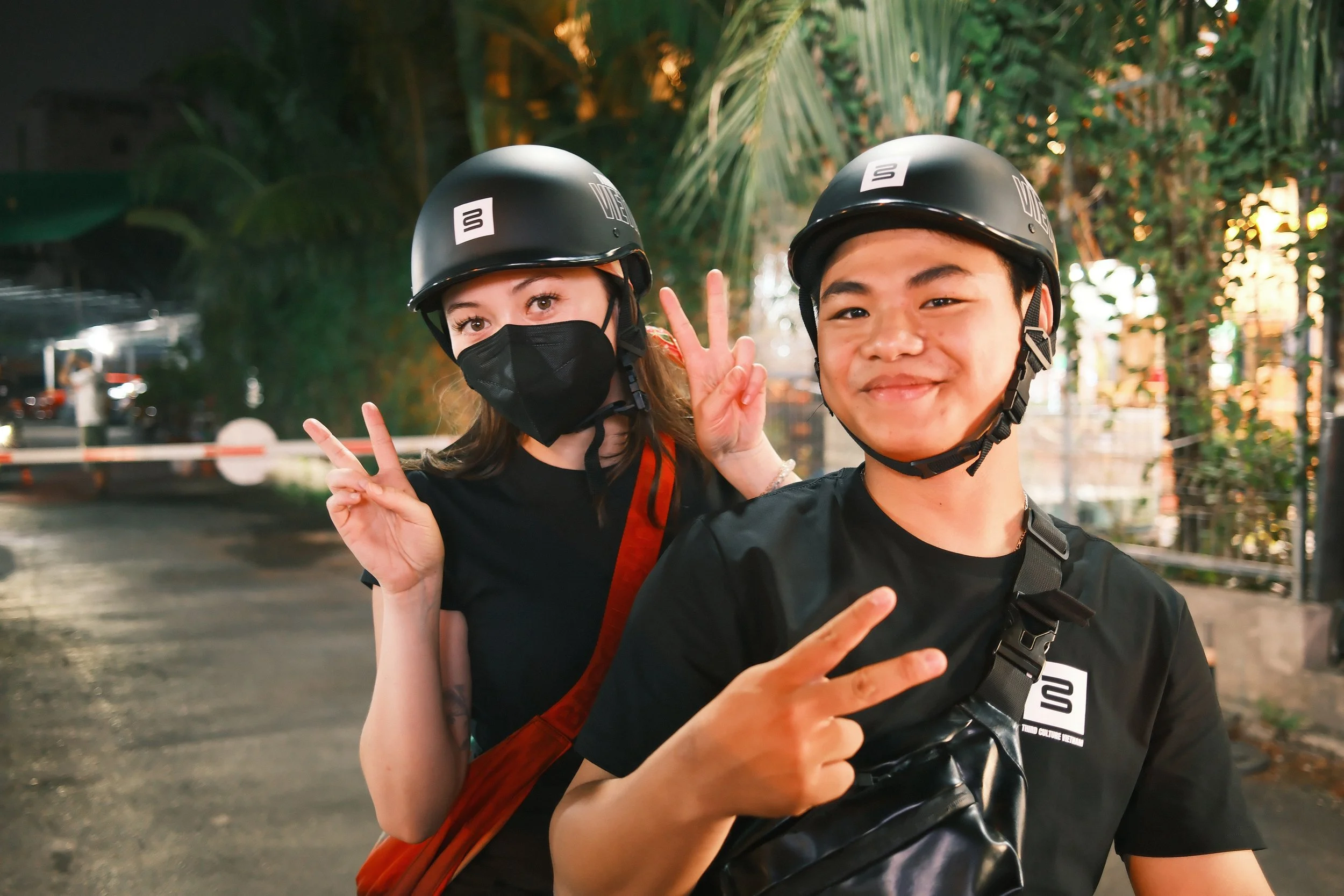 Two young people wearing black helmets and black shirts, making peace signs with their hands, outdoors at dusk.
