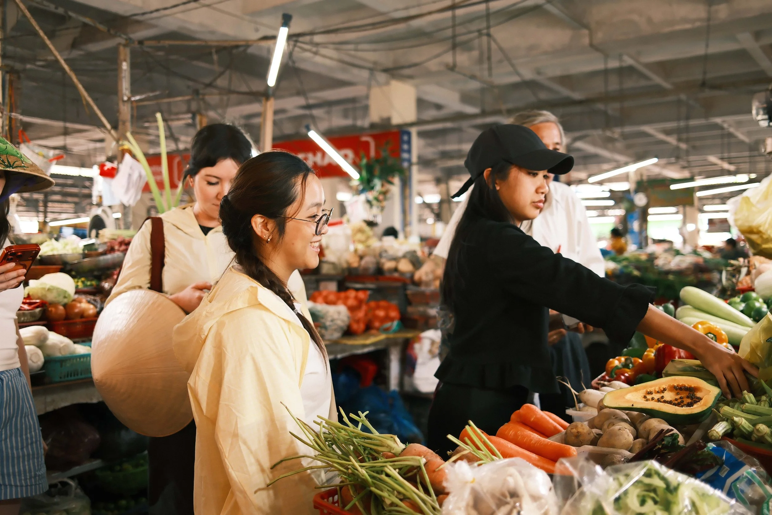 People shopping for produce at a market, looking at vegetables like carrots, onions, and peppers.