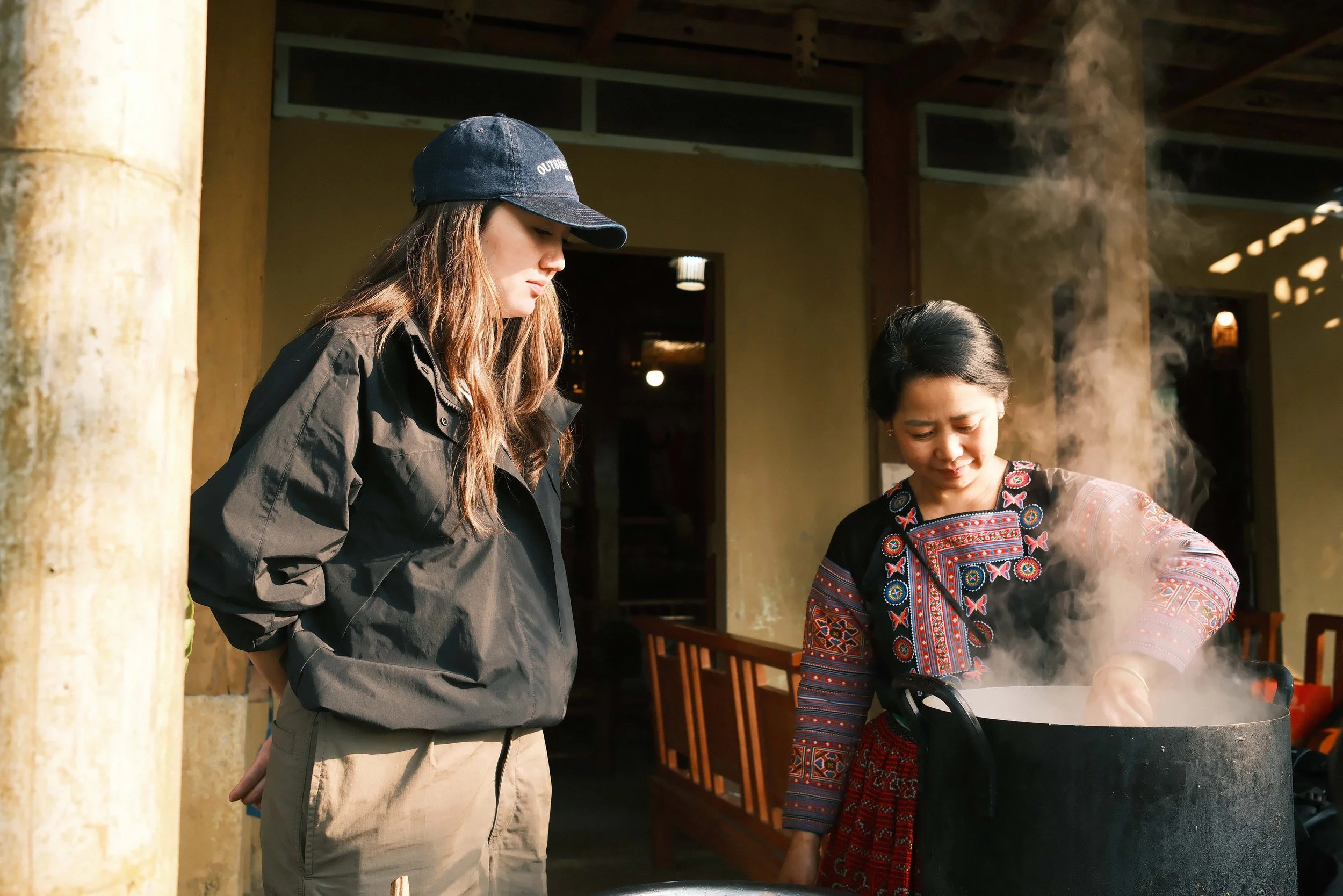 Two women, one young and one older, are cooking outdoors over a grill with steam rising, inside a building with open wood beams.