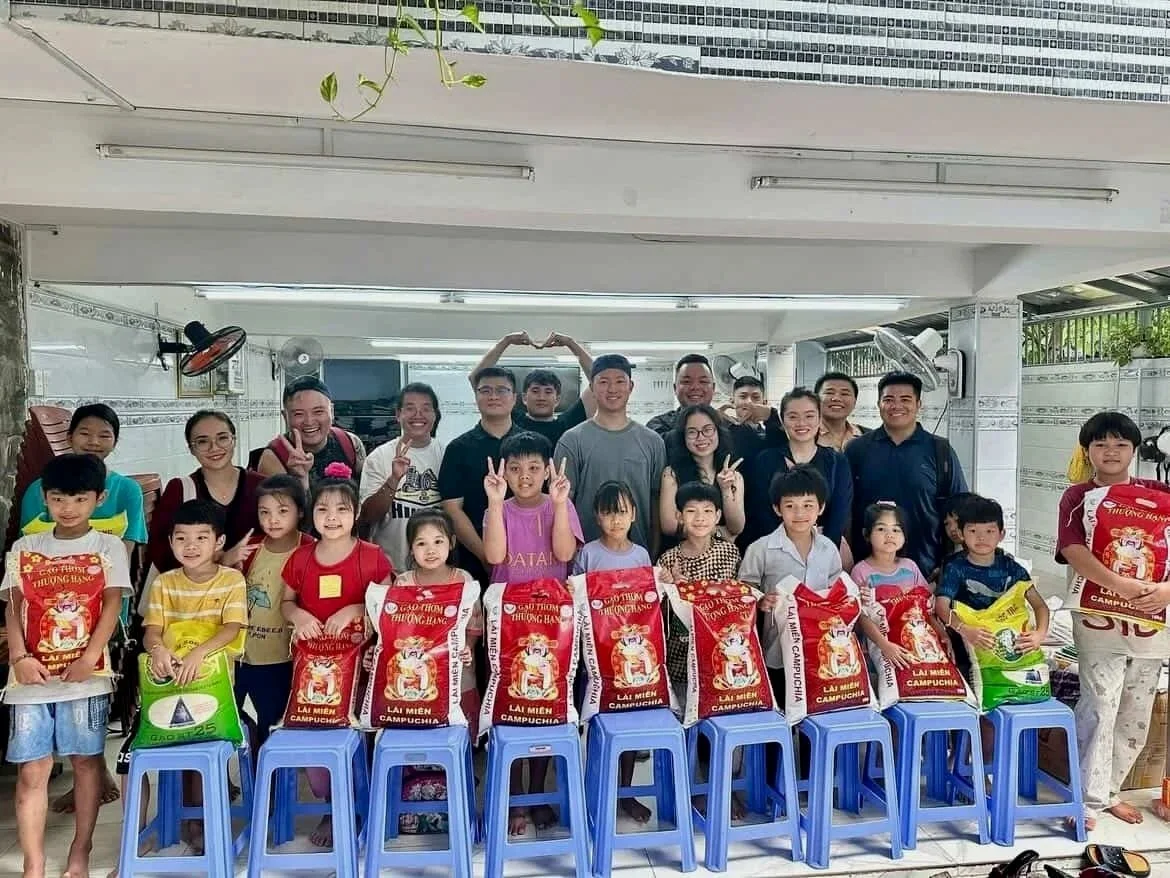 Group of children and adults posing indoors for a photo, with children holding bags of food, in a room with white tiled walls and ceiling fans.
