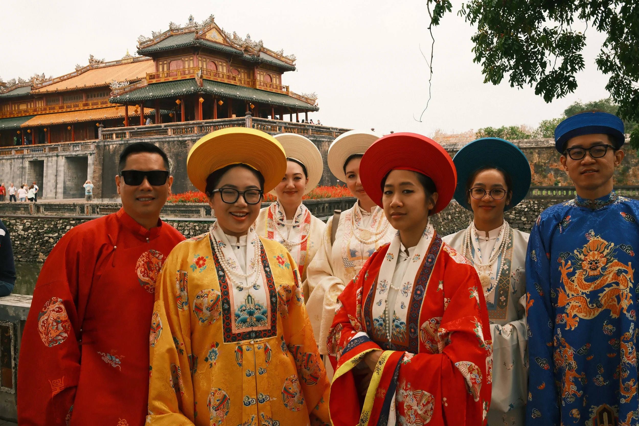 Group of people dressed in traditional Asian clothing, standing outdoors near a historical building with Asian architecture in the background.