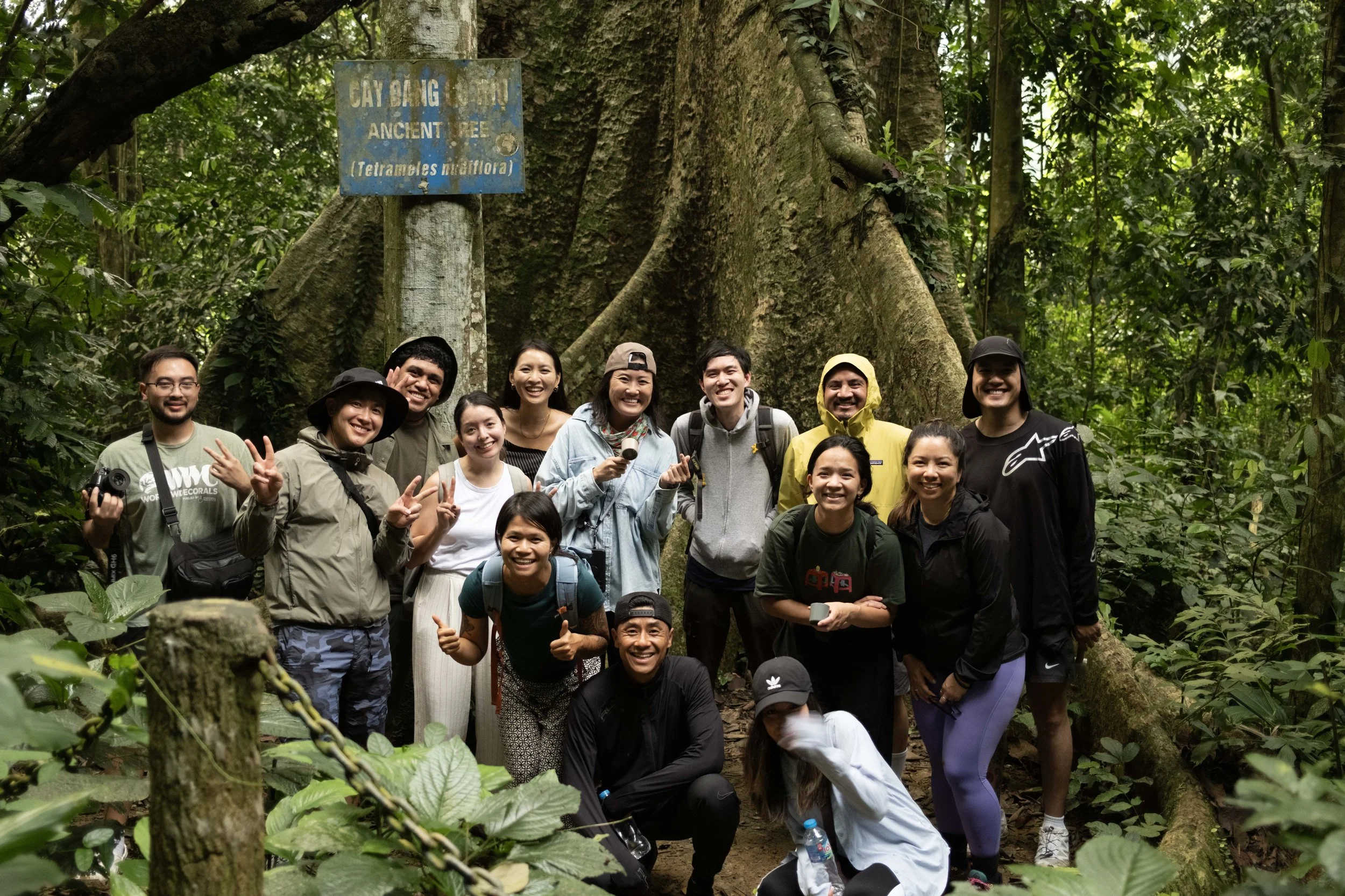 Group of people smiling and posing for a photo in a lush, green forest, with a large tree and a blue sign that reads 'CAYANG DENGU ANCIENT TREE (Tetrameles Nudiiflora)' in the background.