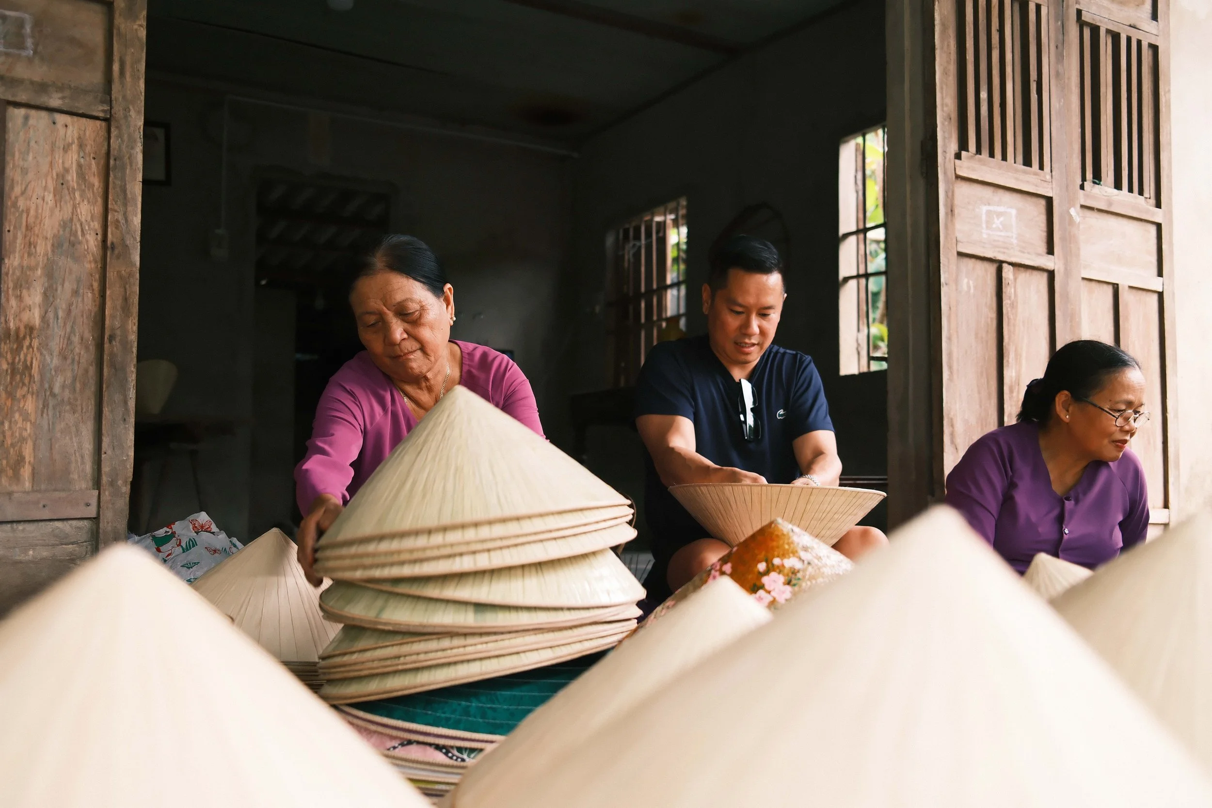 Three women and a young man working on Vietnamese conical hats inside a traditional wooden structure with open windows.
