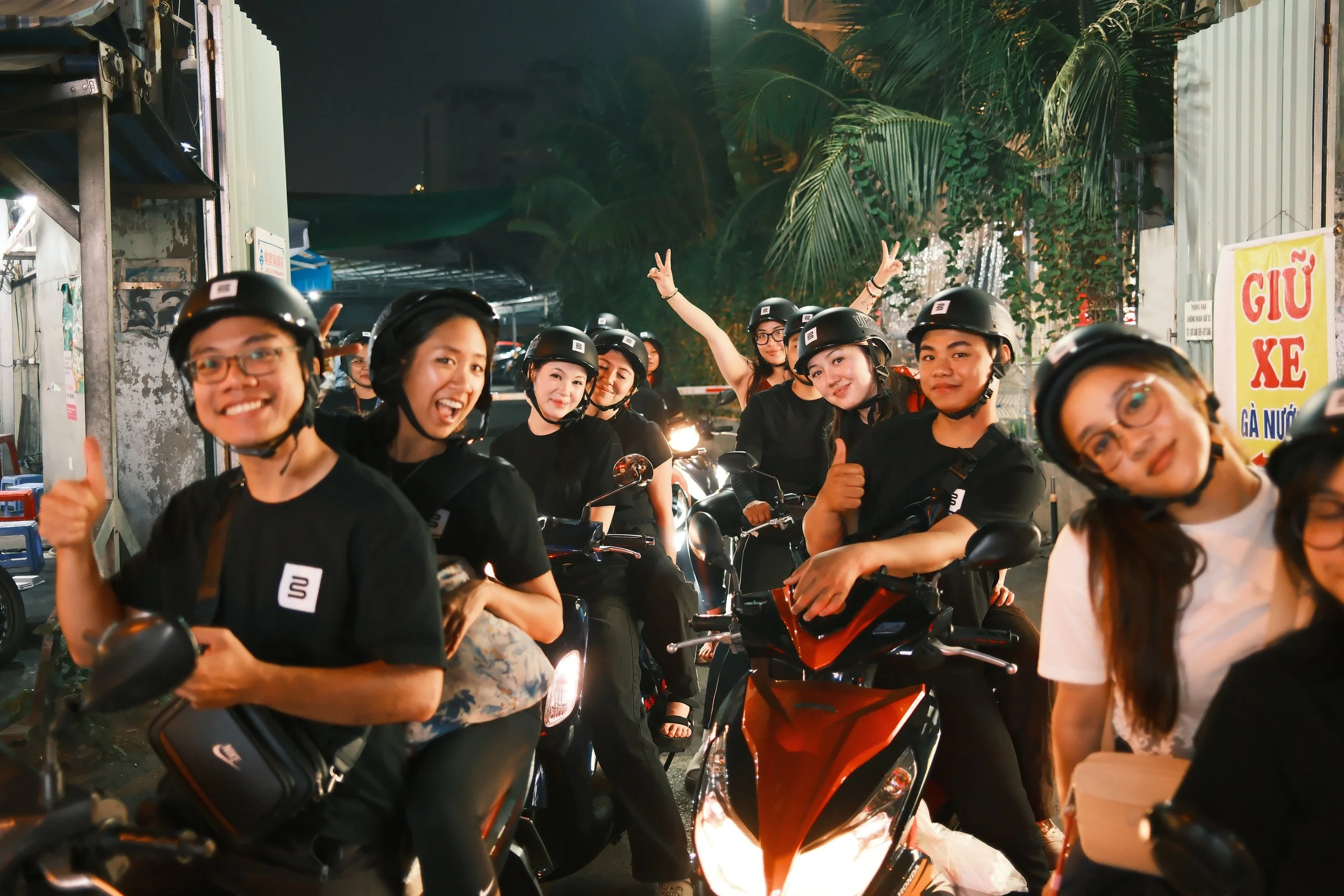 Group of young people in black shirts and helmets gathered on motorbikes at night, smiling and giving thumbs up.