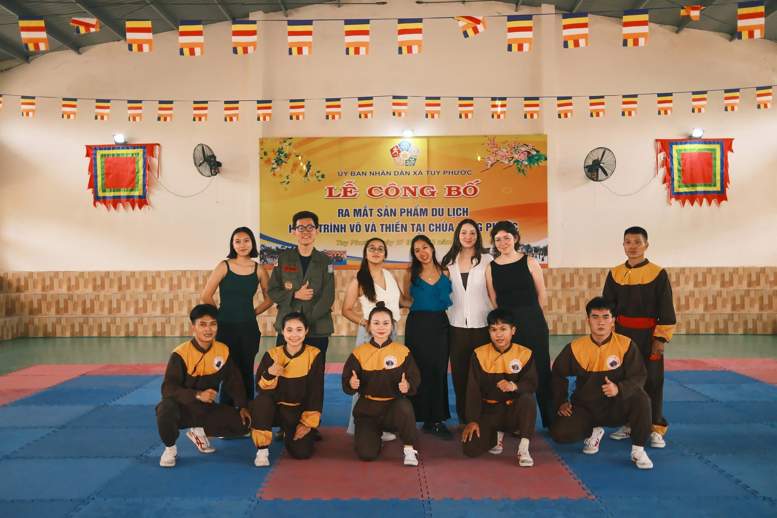 A group of martial artists and officials posing inside a gymnasium decorated with flags and banners, during a martial arts event.