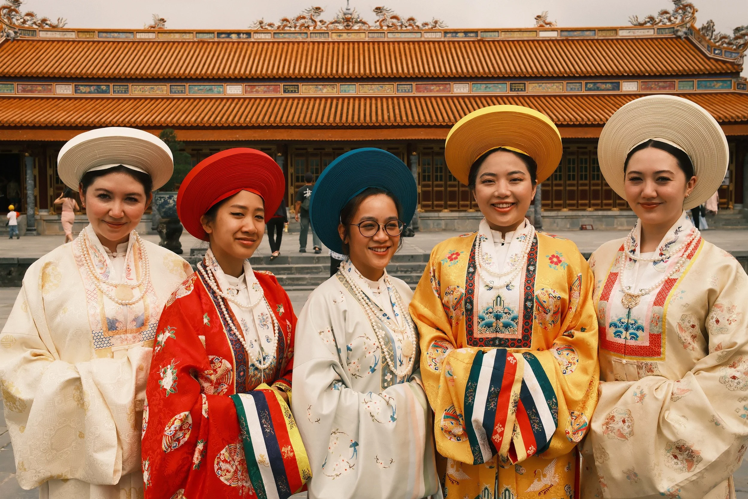 Five women standing in front of a traditional Asian building, wearing colorful traditional dresses and wide-brimmed hats.