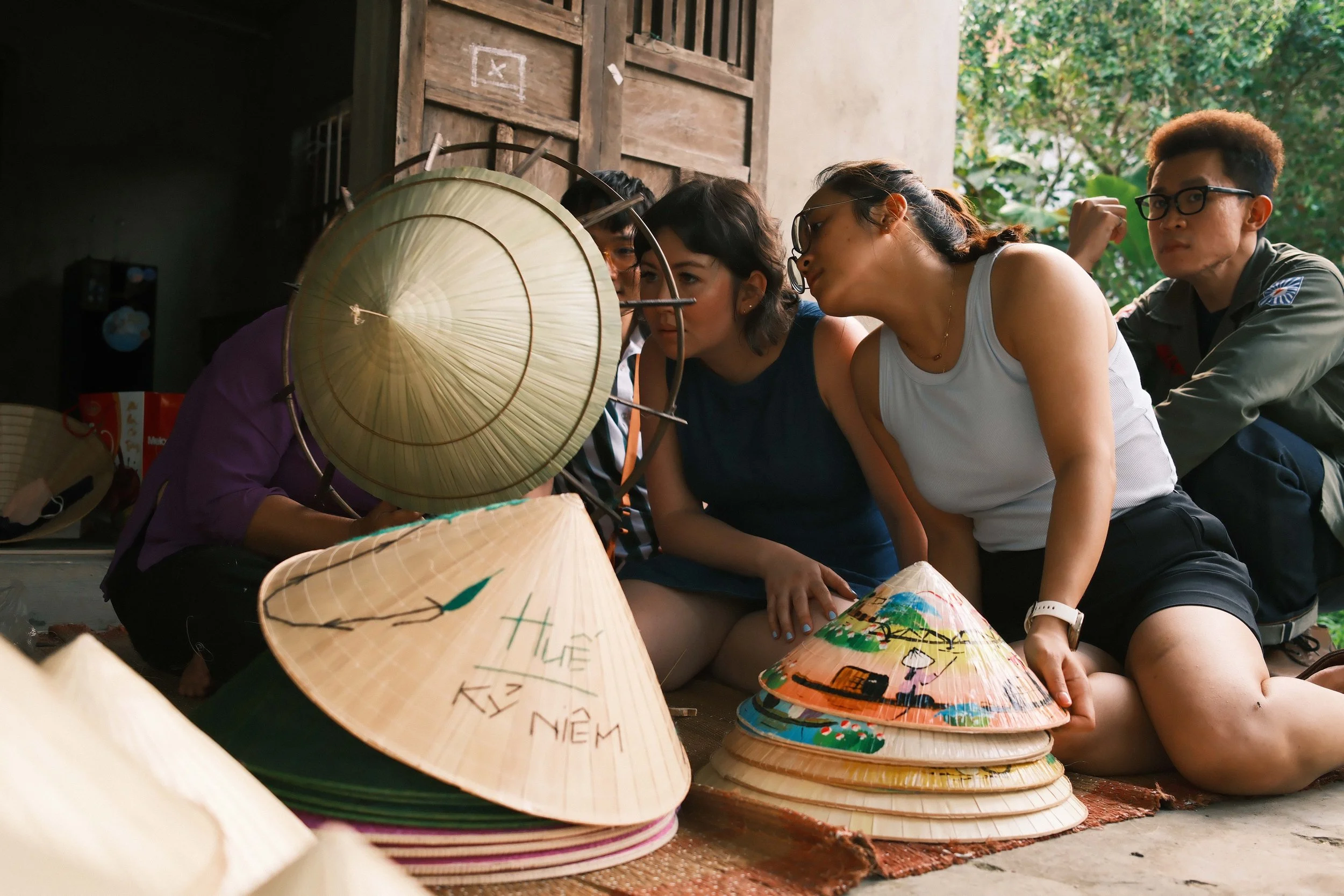 Group of people sitting on the ground indoors, closely examining traditional Asian conical hats called non la, with some hats decorated with writings and drawings, in a room with wooden walls and open window showing greenery outside.