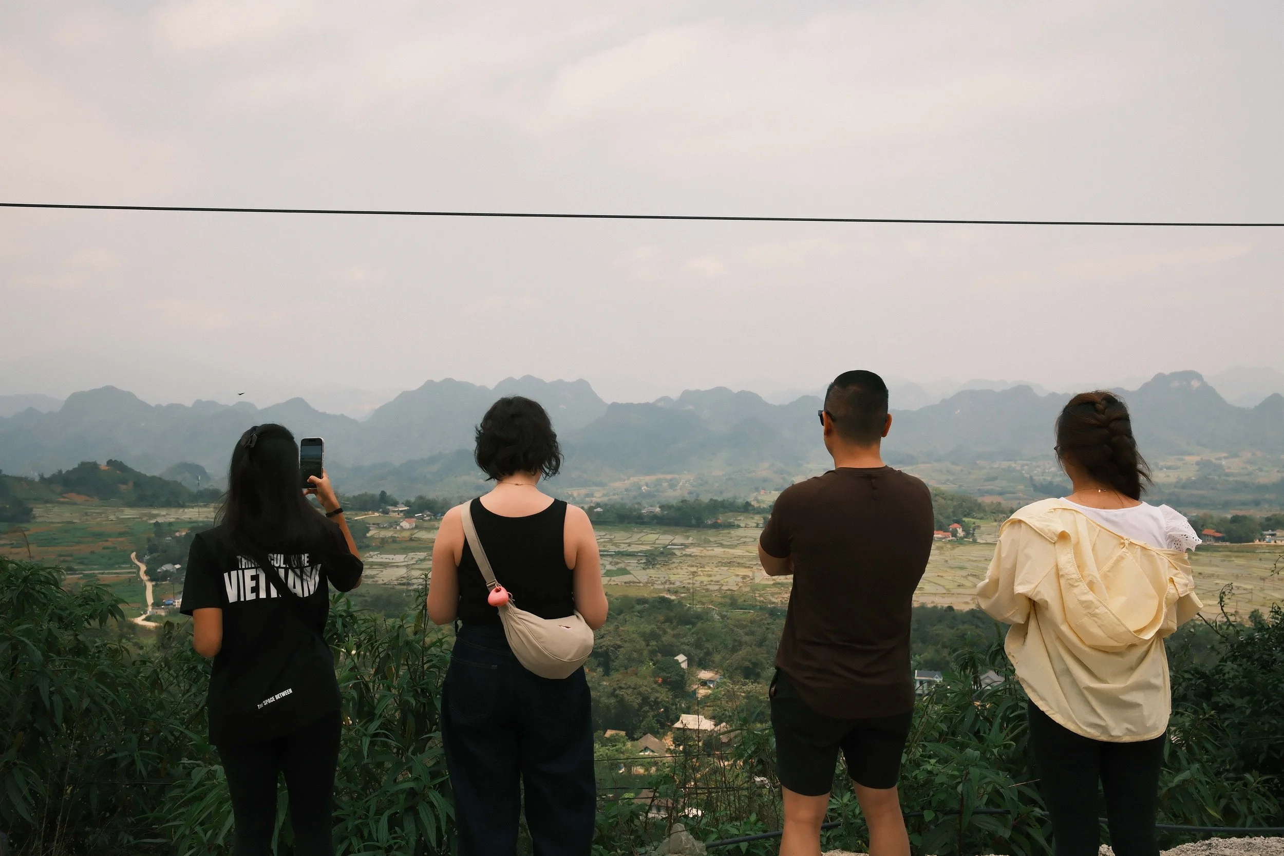 Four people standing on a hill overlooking a valley with mountains in the background, one person is taking a photo with a smartphone.