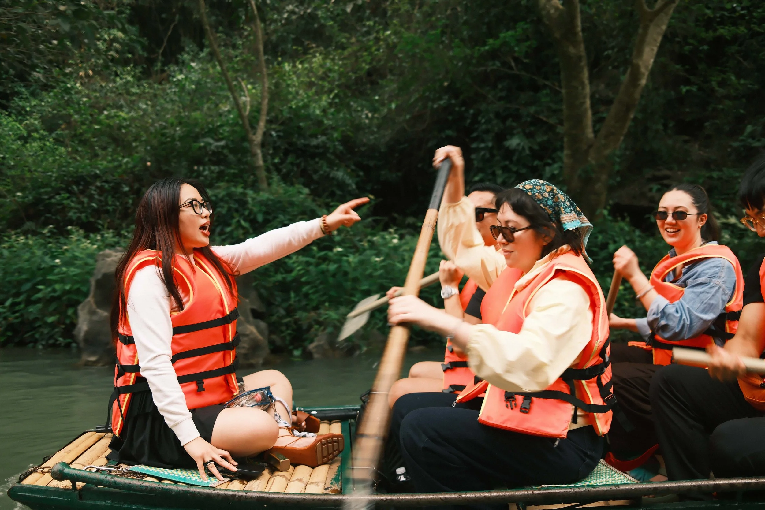 Group of women in life jackets sitting on a bamboo raft paddling in a river surrounded by lush green trees.