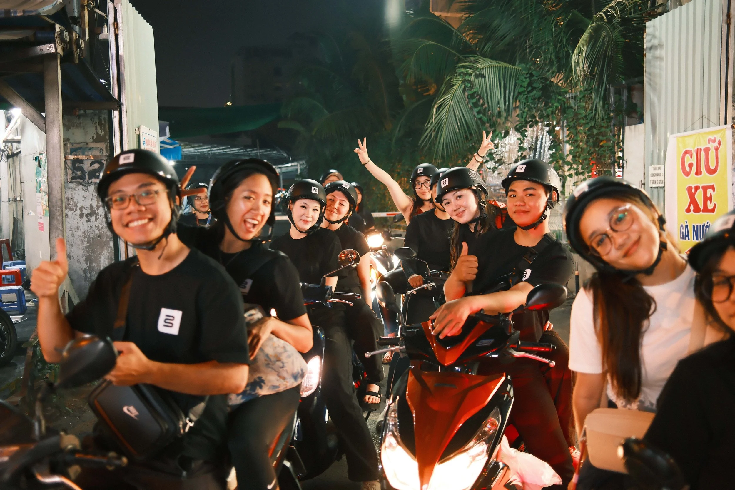 Group of smiling people wearing helmets and sitting on motorbikes in an outdoor night market area.