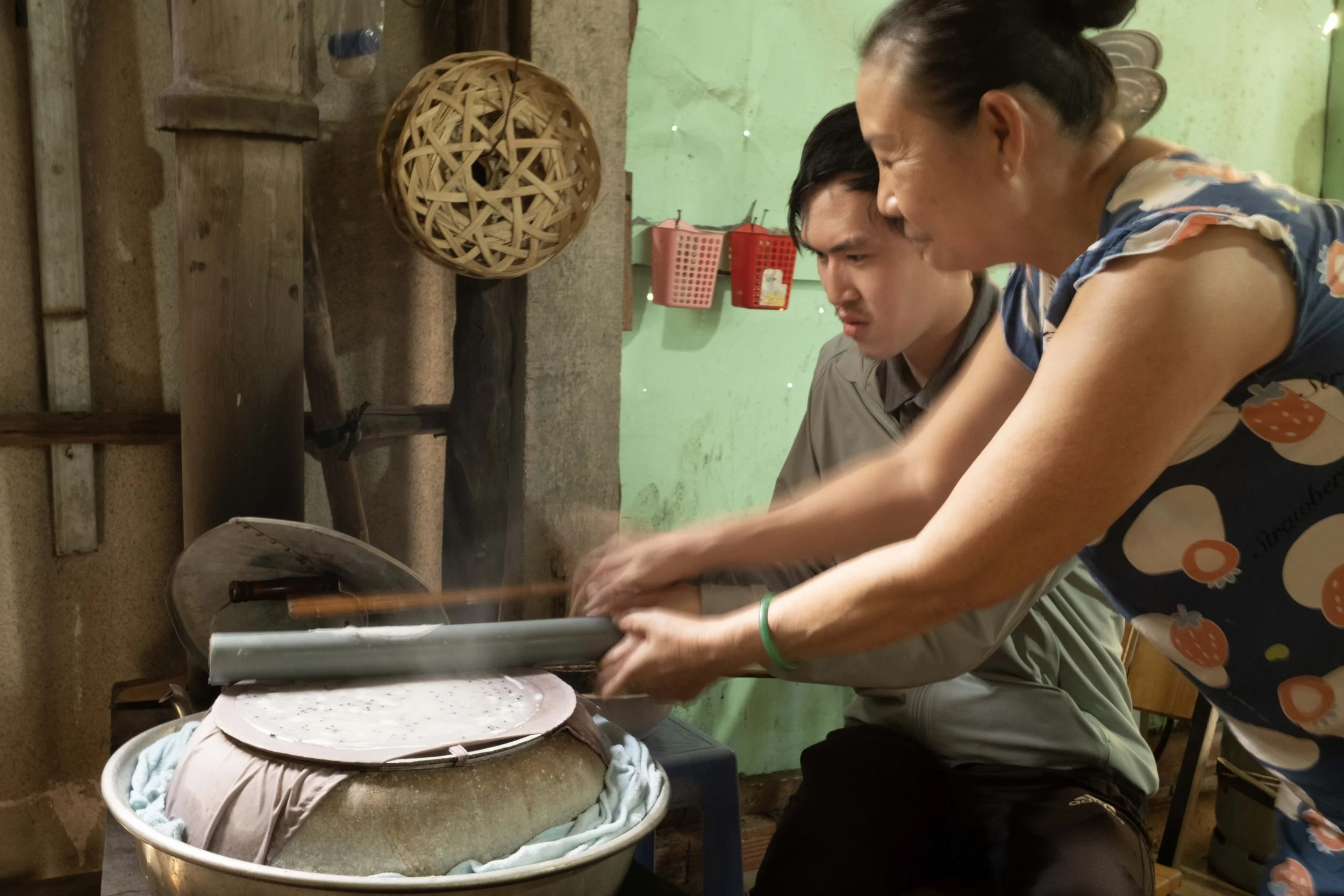 Two women are preparing dough or food together in a rustic kitchen with a green wall, a wooden work surface, and a large metal mixing bowl, with two men observing.