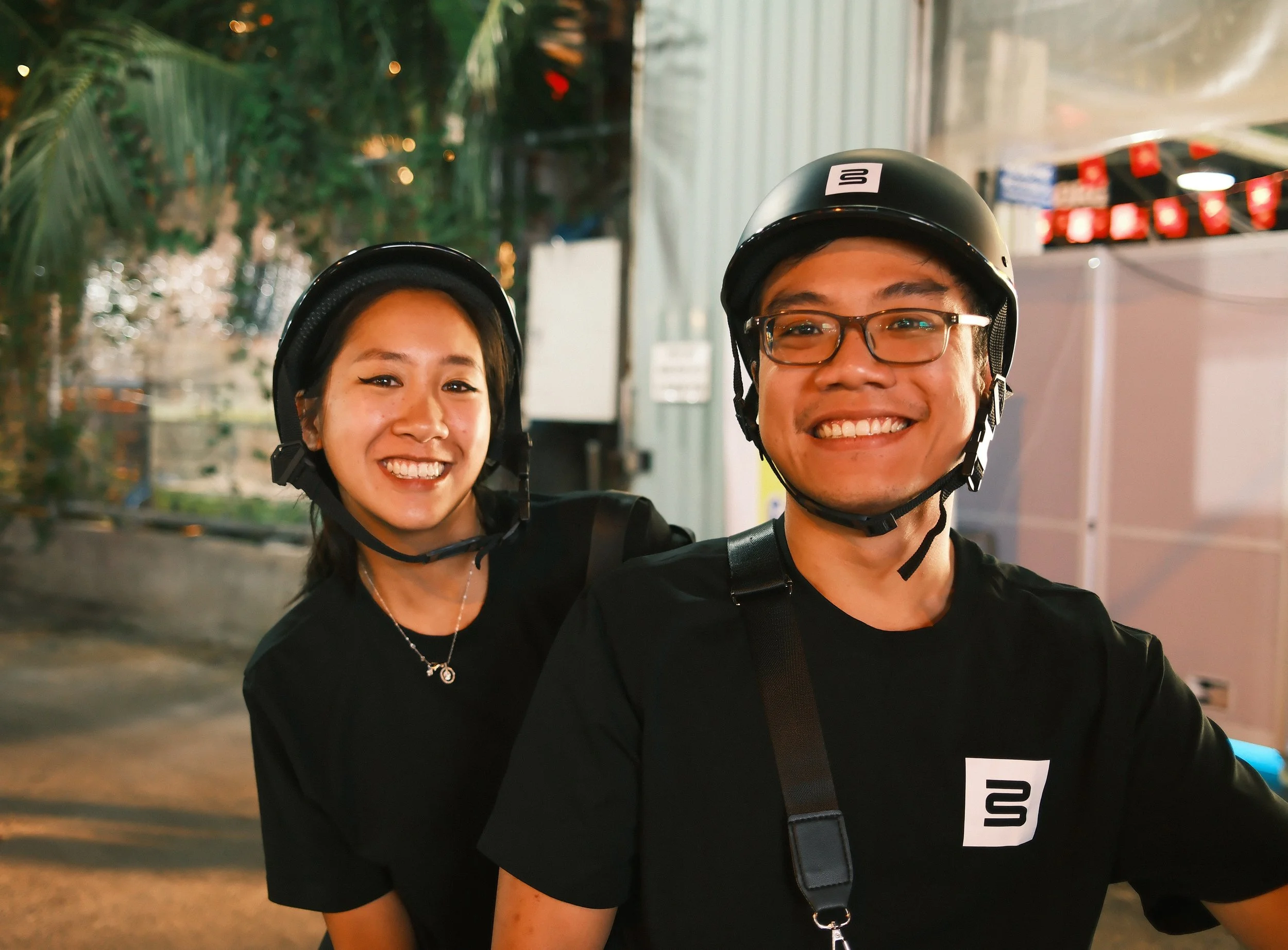 A smiling young man and woman wearing black T-shirts and helmets, standing outdoors at night.