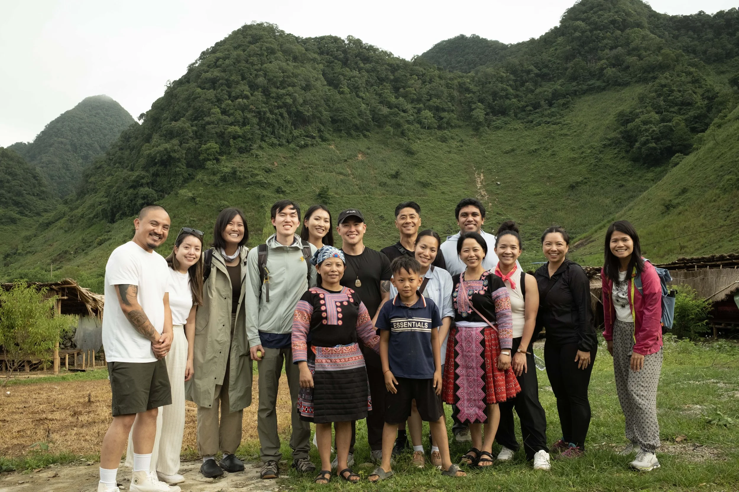 Group of diverse people standing outdoors in front of green mountains, smiling for a photo.