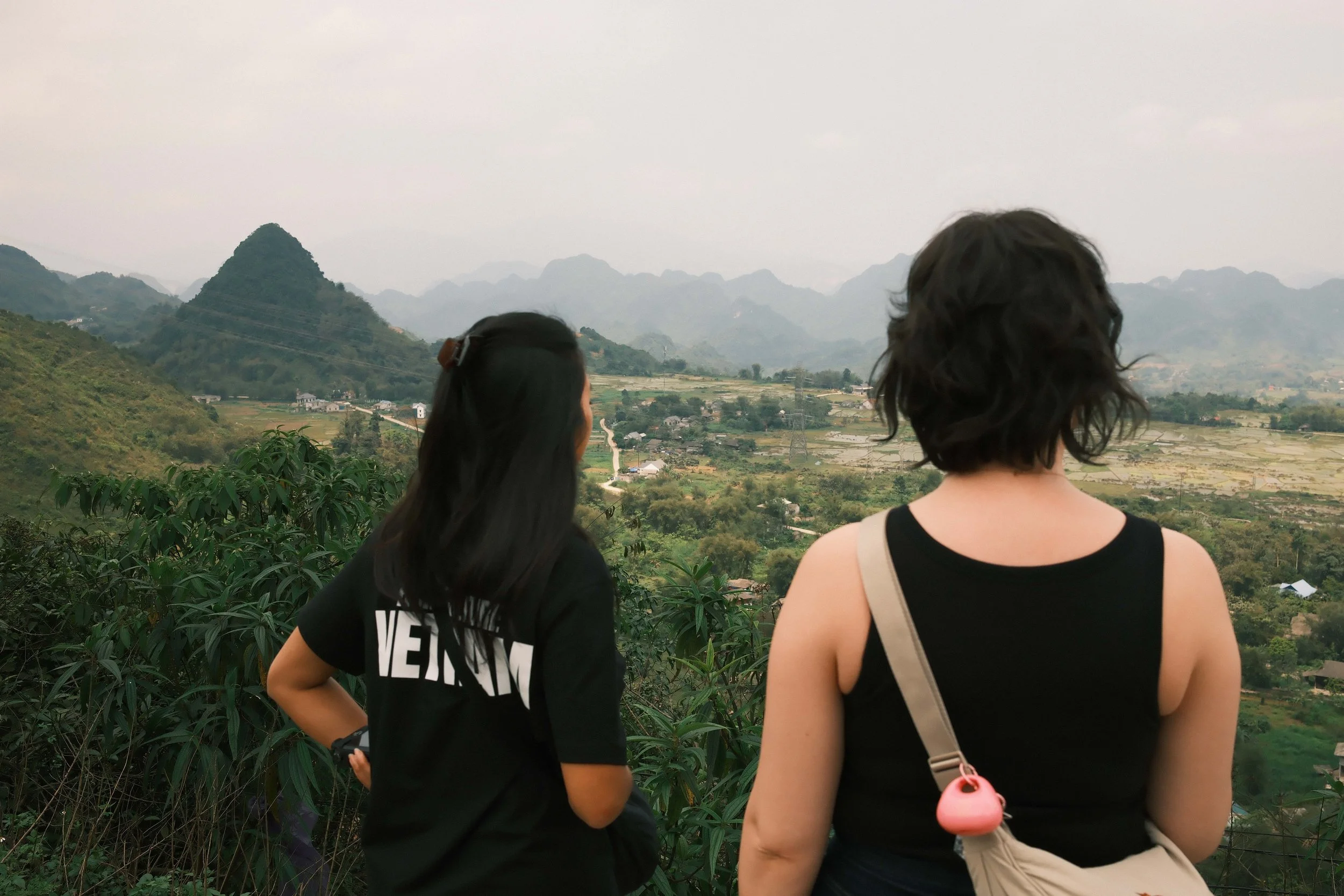 Two women with dark hair standing outdoors, looking at a mountainous landscape with overlapping green hills and small houses in the valley.
