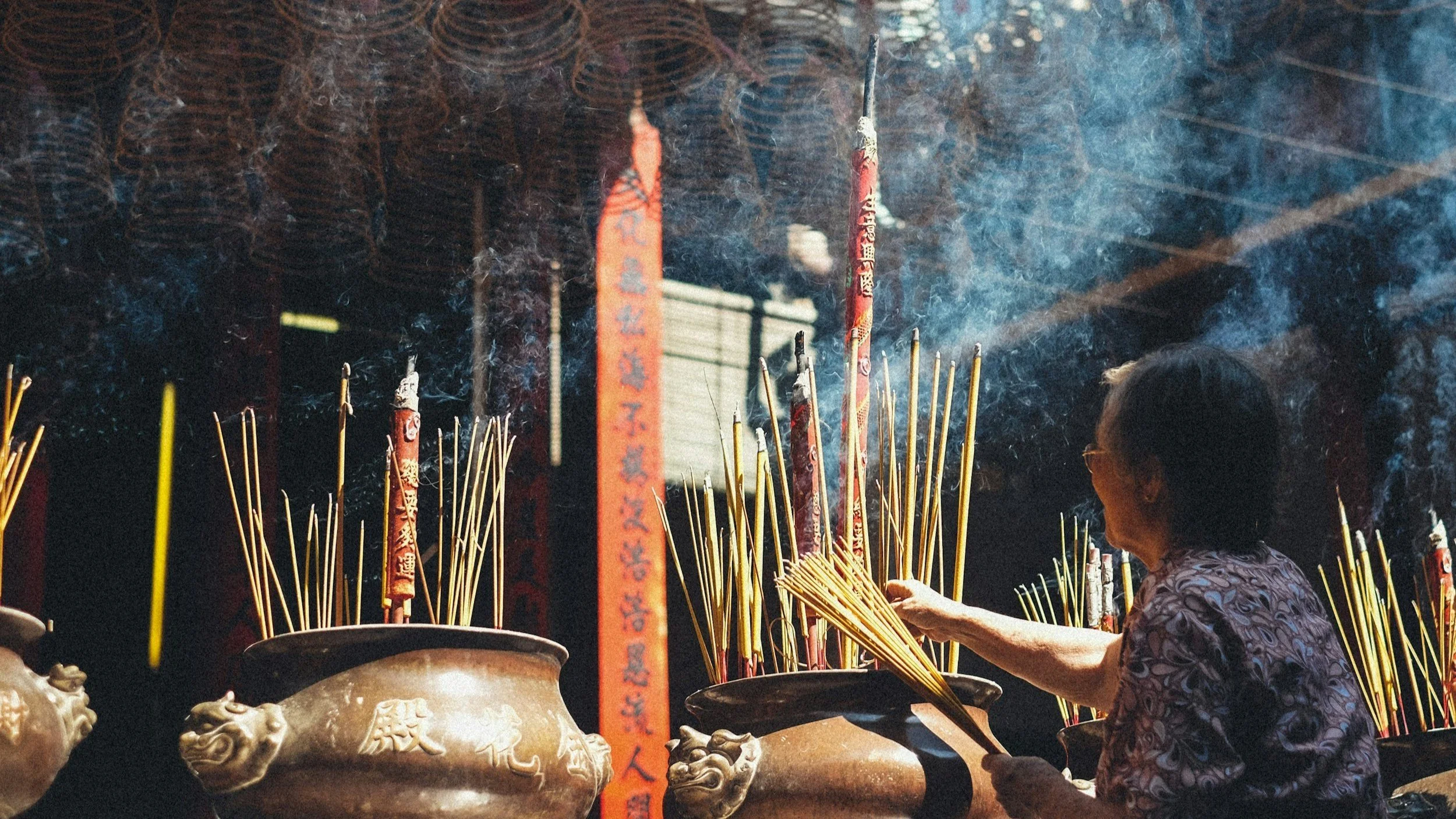 A woman lighting incense sticks at a temple, with smoke rising from the incense, large bronze urns, and red prayer banners with Chinese characters.