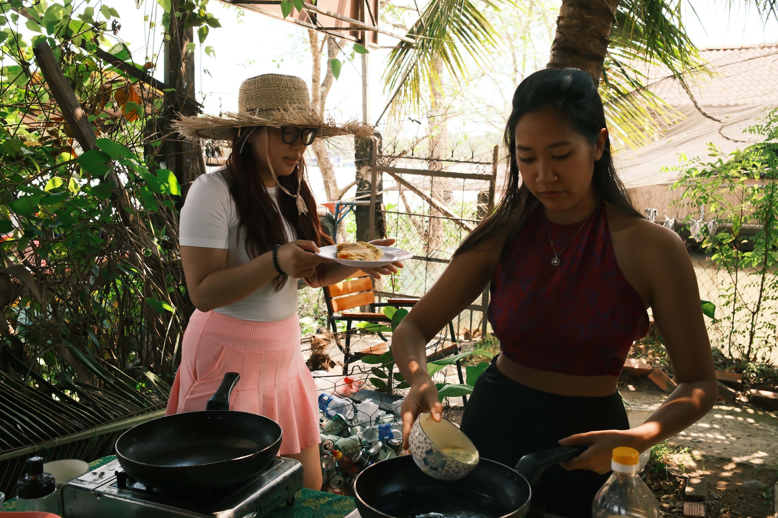 Two women stand outdoors among green foliage and trees, cooking on portable stoves. One woman is wearing a wide-brimmed straw hat and pink skirt, holding a plate, while the other woman is wearing a sleeveless top, focused on cooking.