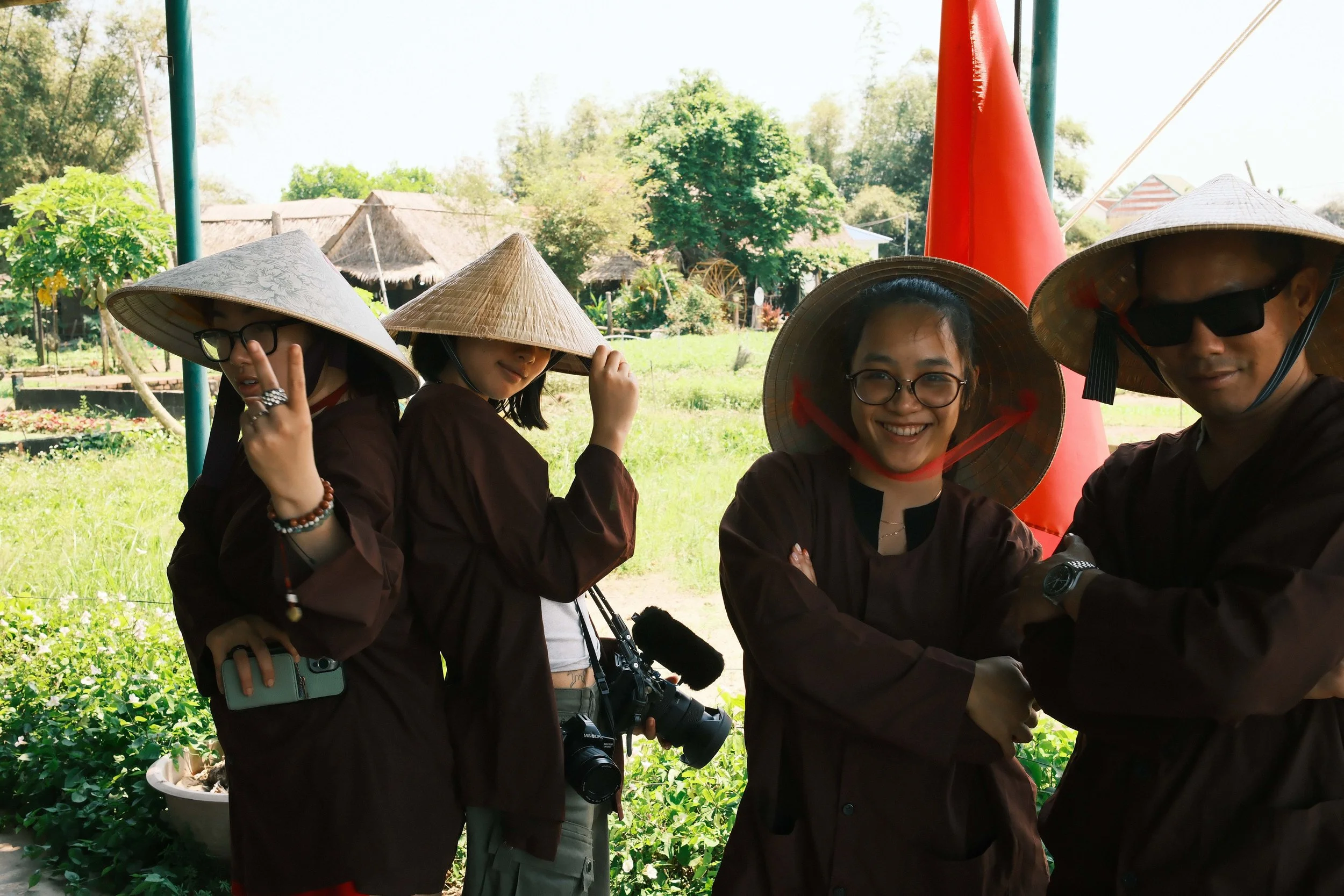 Group of four people outdoors wearing traditional Asian conical hats, some with glasses, smiling and posing with camera equipment, in a lush green garden setting.