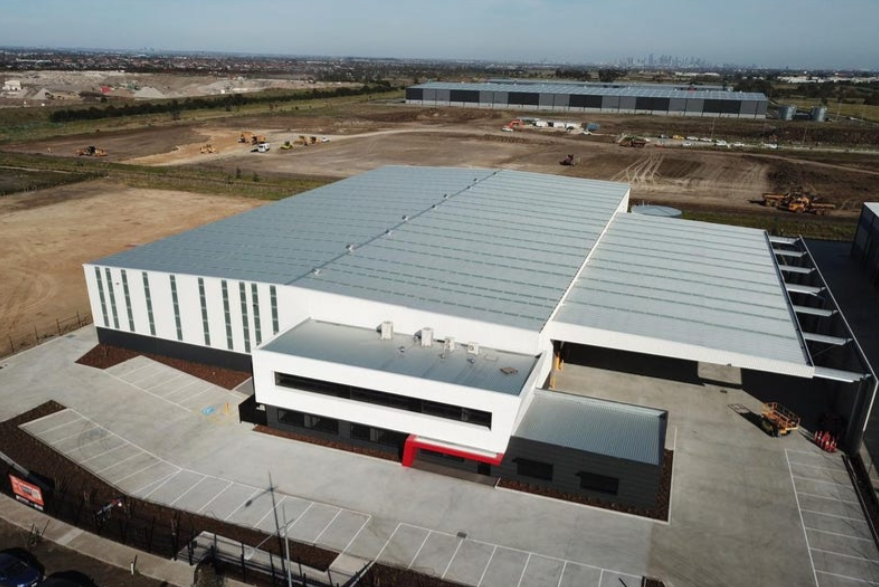 A large industrial warehouse building with a white exterior and metal roof, surrounded by a parking lot, under construction with some machinery nearby, in an area with open land and distant city skyline.