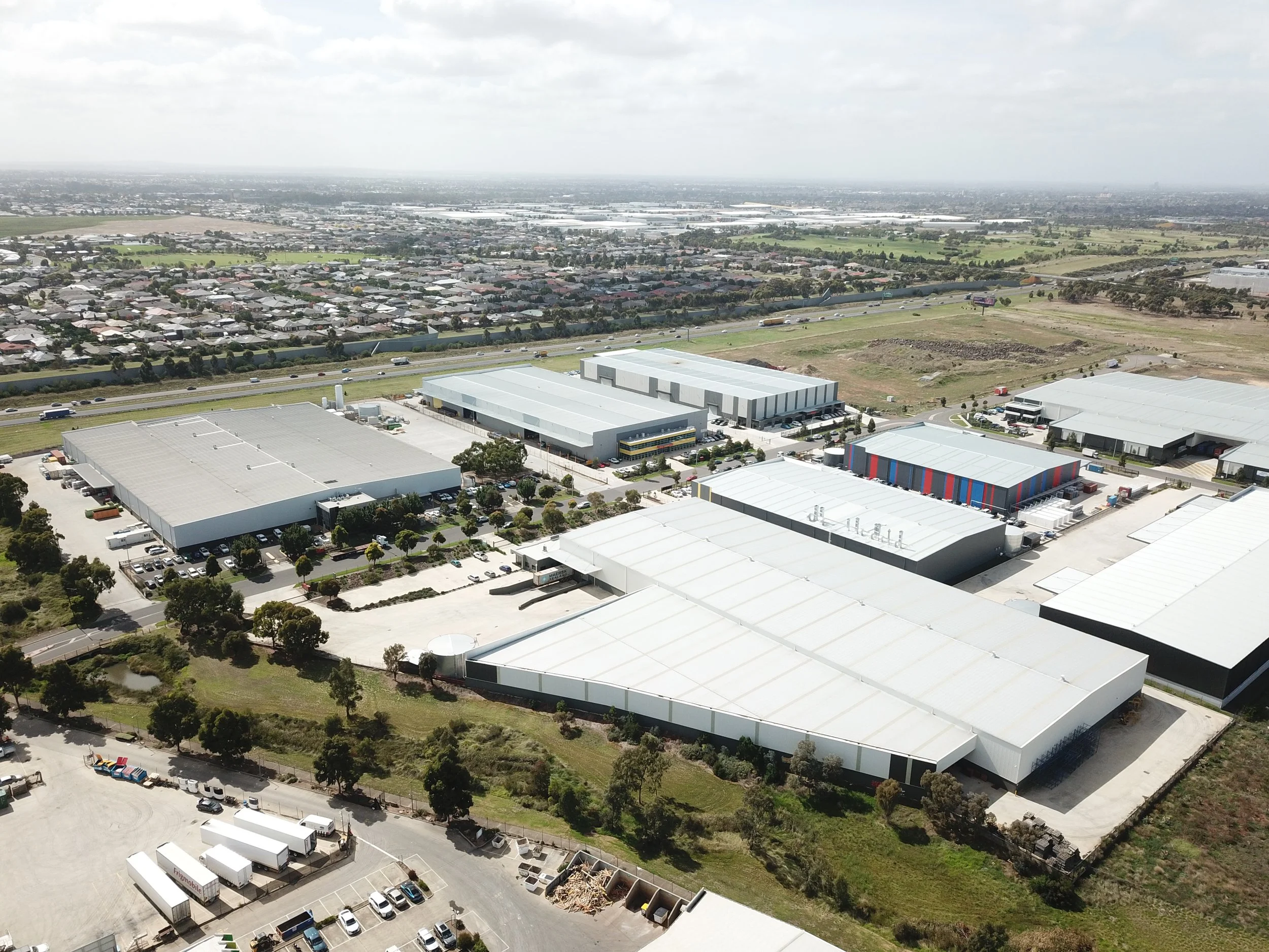 Aerial view of an industrial warehouse complex with multiple large buildings, parking lots, and nearby residential area under partly cloudy skies.