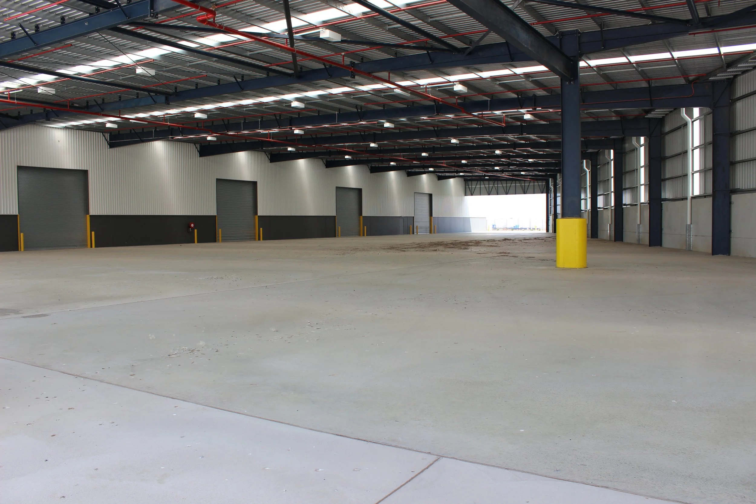 Empty warehouse with large open floor space, multiple roller doors along the wall, metal roof, steel support beams, and windows on the side wall.