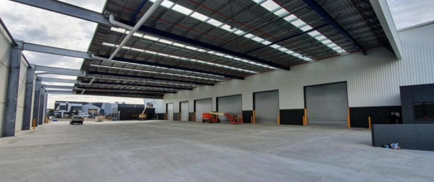 View of a large industrial warehouse with a wide open loading area, multiple closed loading dock doors, a crane, and construction equipment, under a partially covered roof.