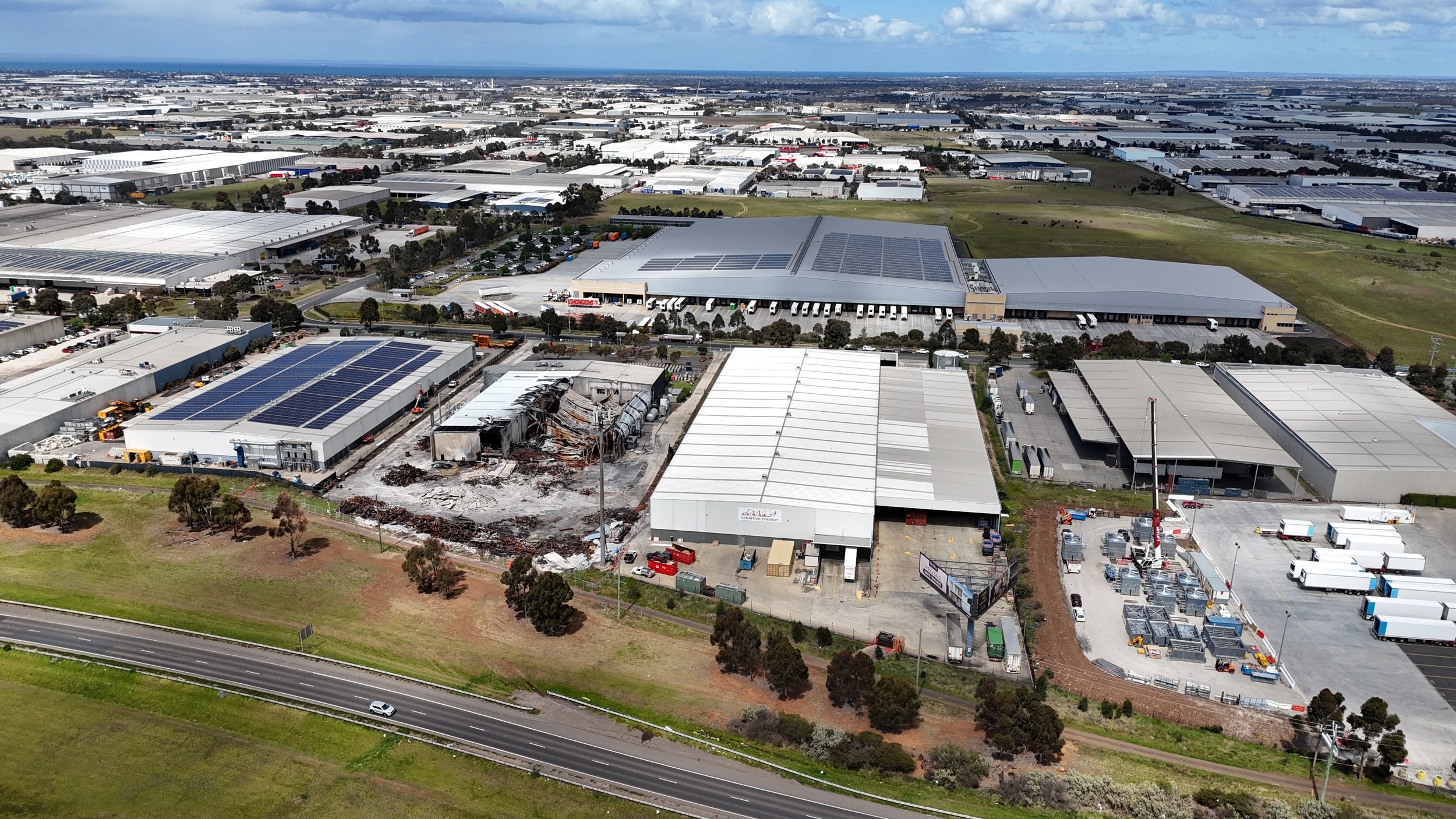 Aerial view of a commercial industrial area with warehouses, some with solar panels, and construction ongoing on one building, with trucks and cars parked around.