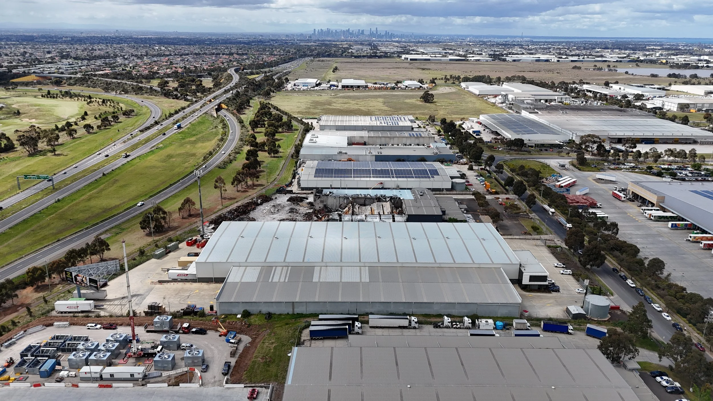 Aerial view of a commercial or industrial area with warehouses, trucks, and a highway with cars, with urban skyline in the distance under partly cloudy sky.