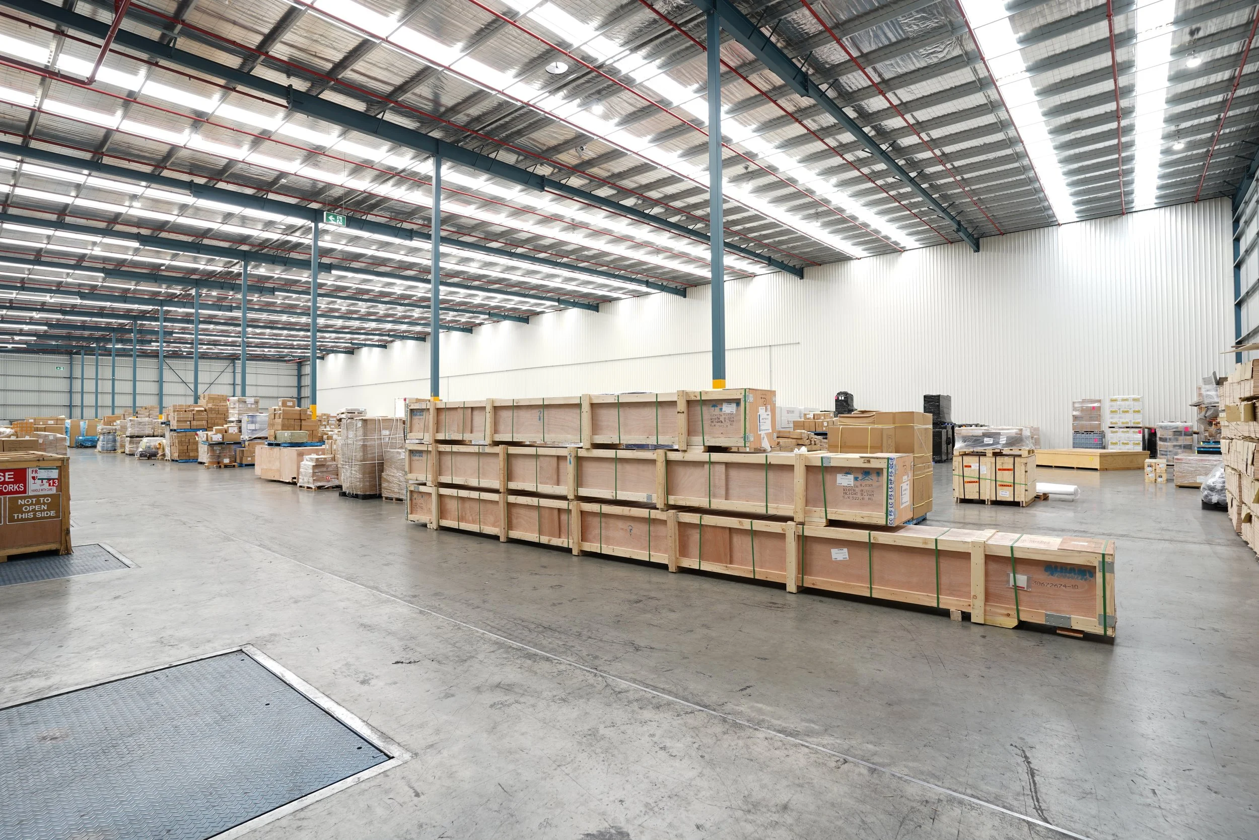 A spacious warehouse with high ceiling, metal support beams, and ray-panel lighting, filled with stacks of cardboard boxes and wooden crates on a polished concrete floor.