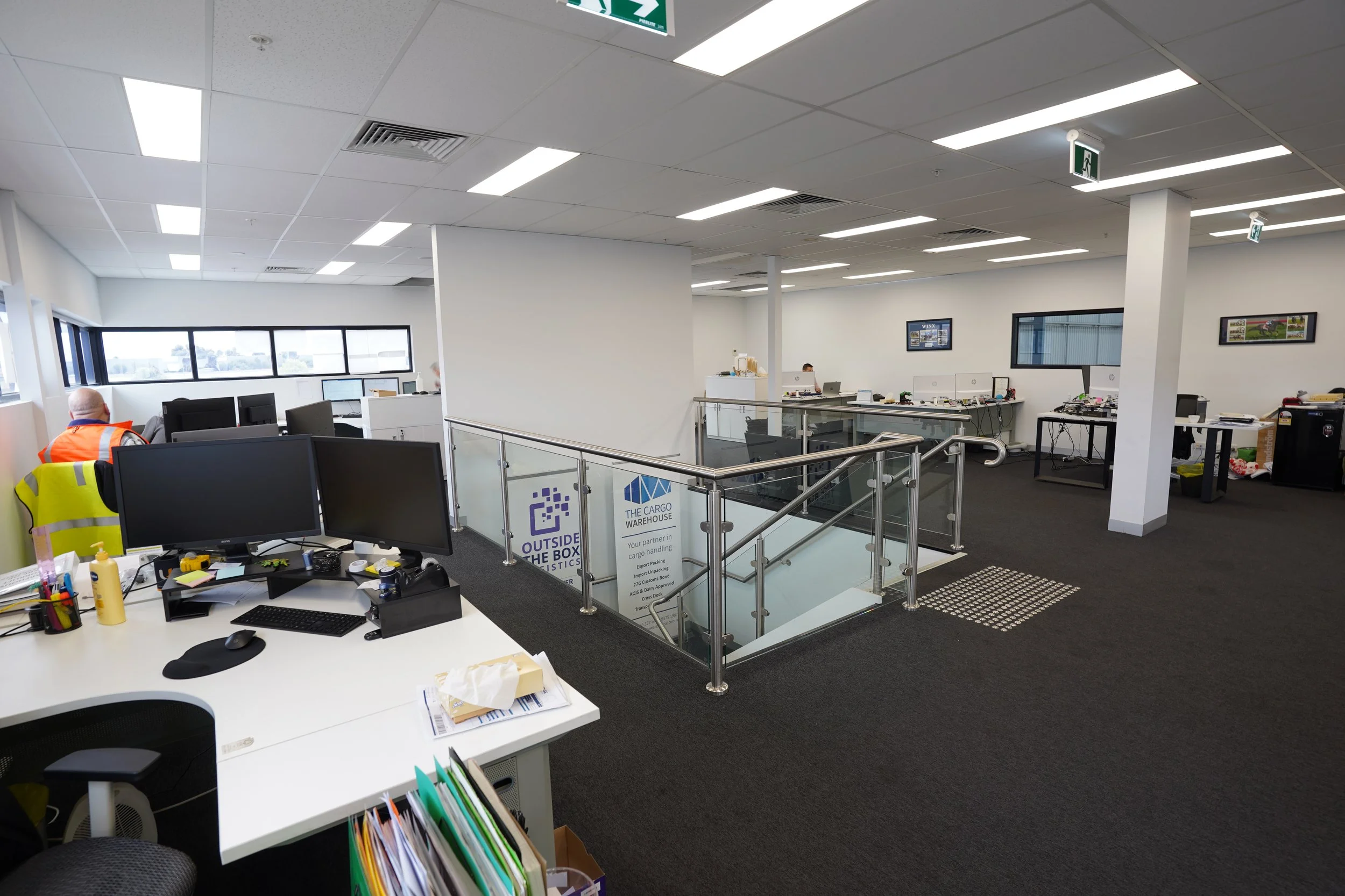View of an office space with desks, computers, and a staircase with glass railings, white walls, and large windows letting in natural light.