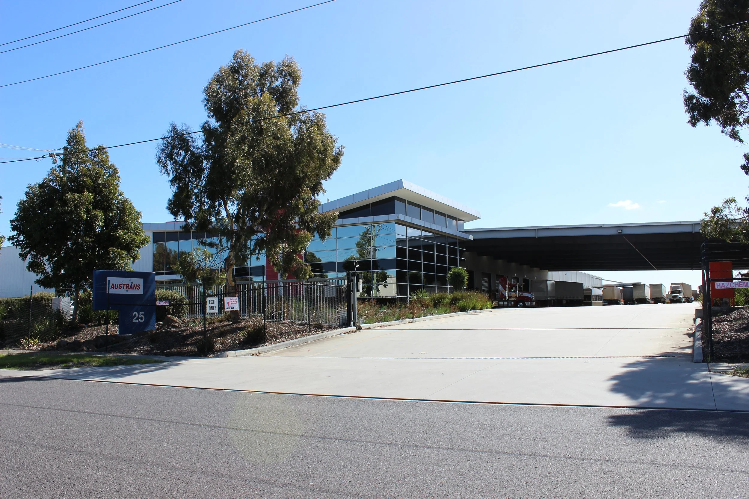 Modern industrial warehouse with a glass exterior and a loading dock with trucks parked, surrounded by trees and a driveway, under a clear blue sky.