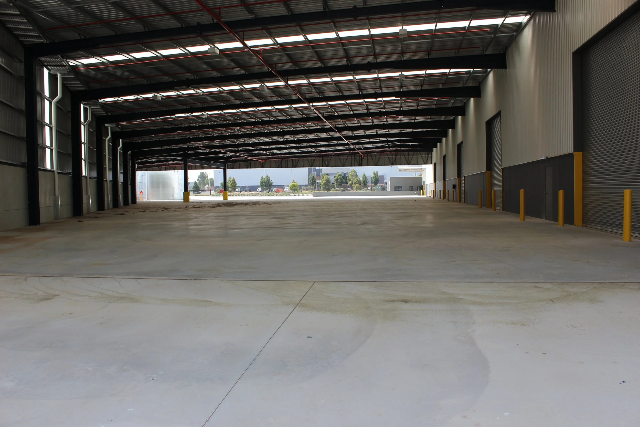 Empty warehouse with large open space, concrete floor, metal walls, and roll-up doors, with outdoor view of trees and buildings