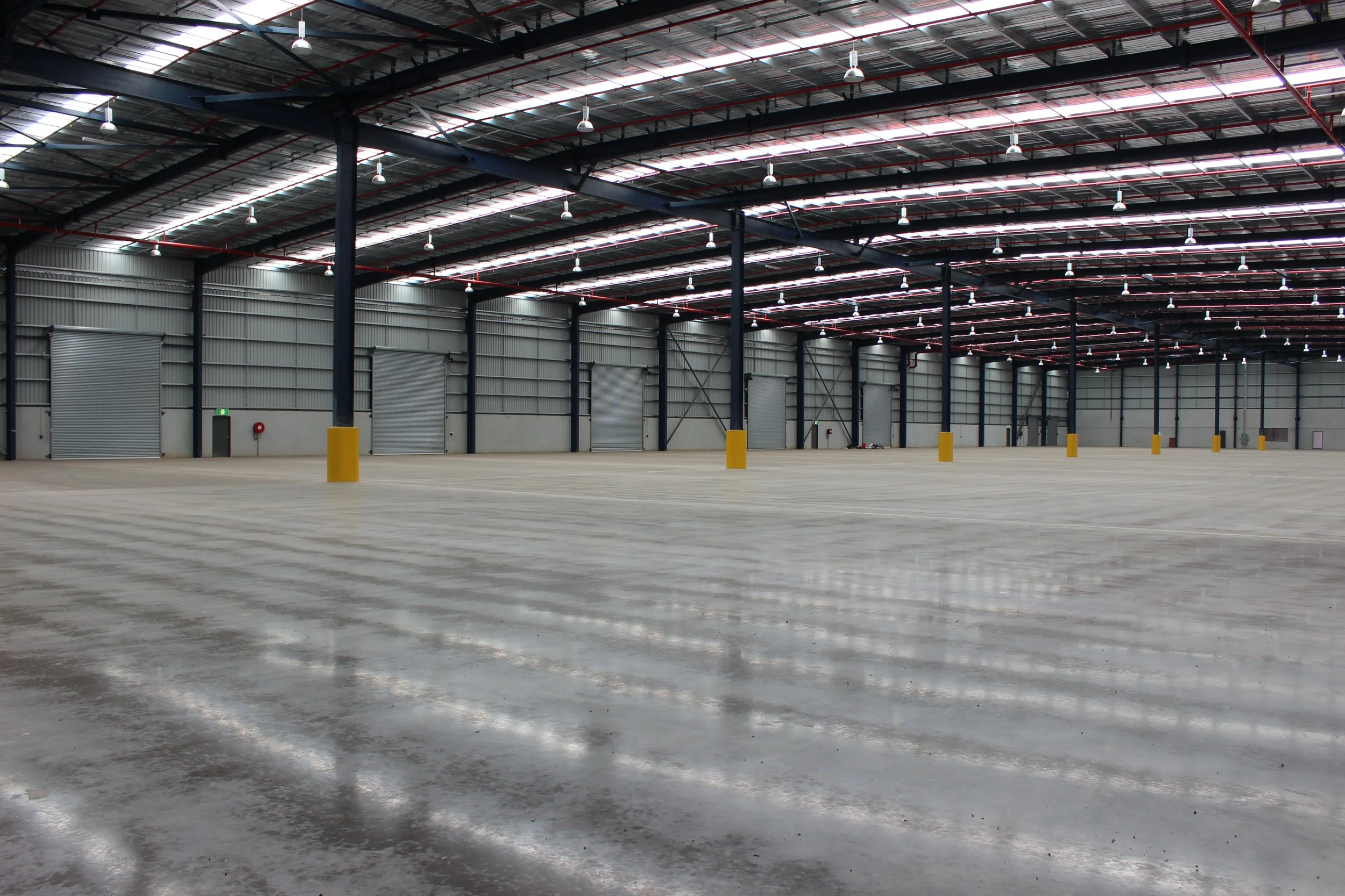 Empty warehouse with high ceilings, metal walls, and large rolling doors, illuminated by ceiling lights.
