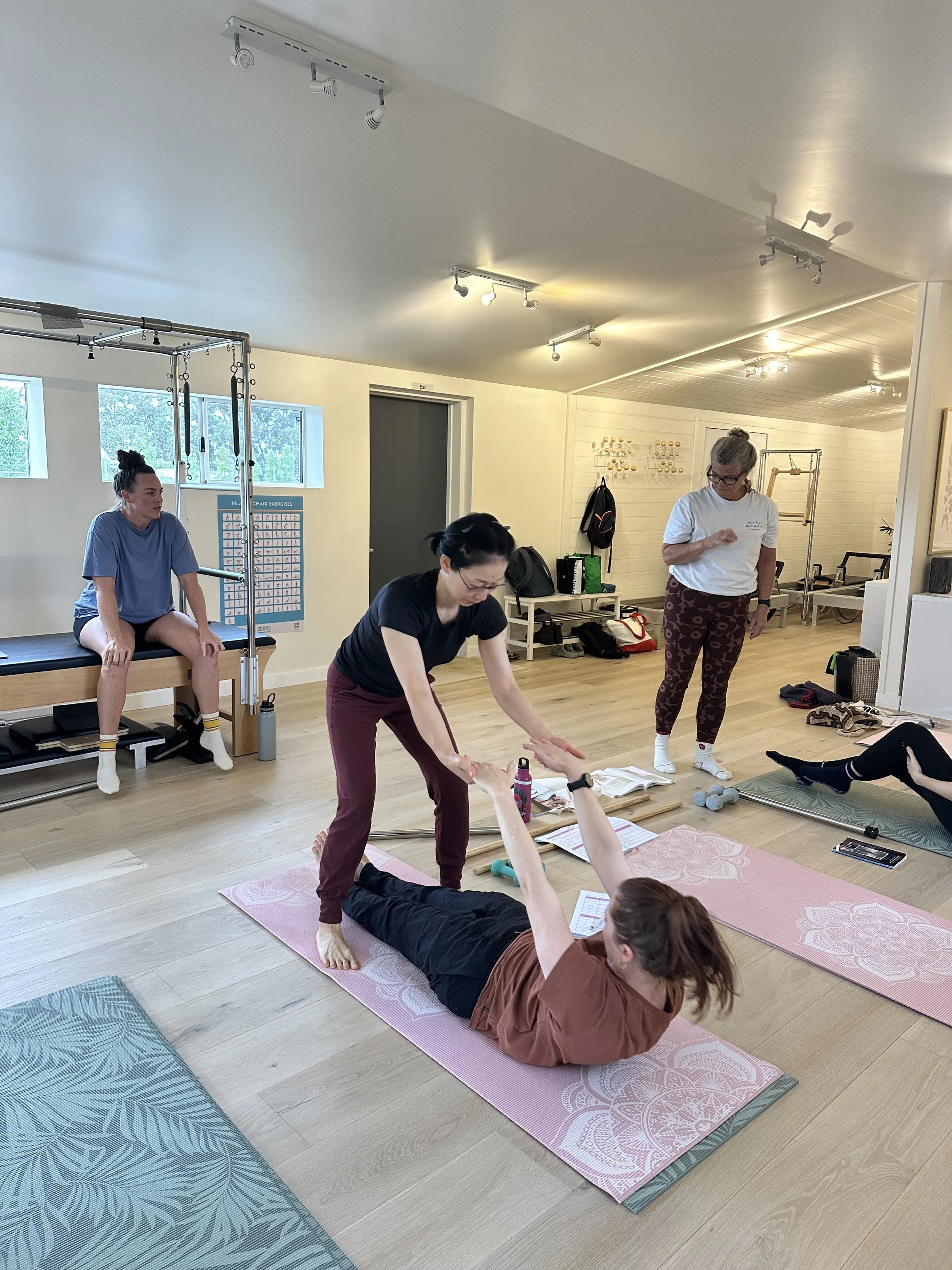 A group Pilates class in a studio with participants doing exercises and an instructor assisting a woman lying on a pink mat.