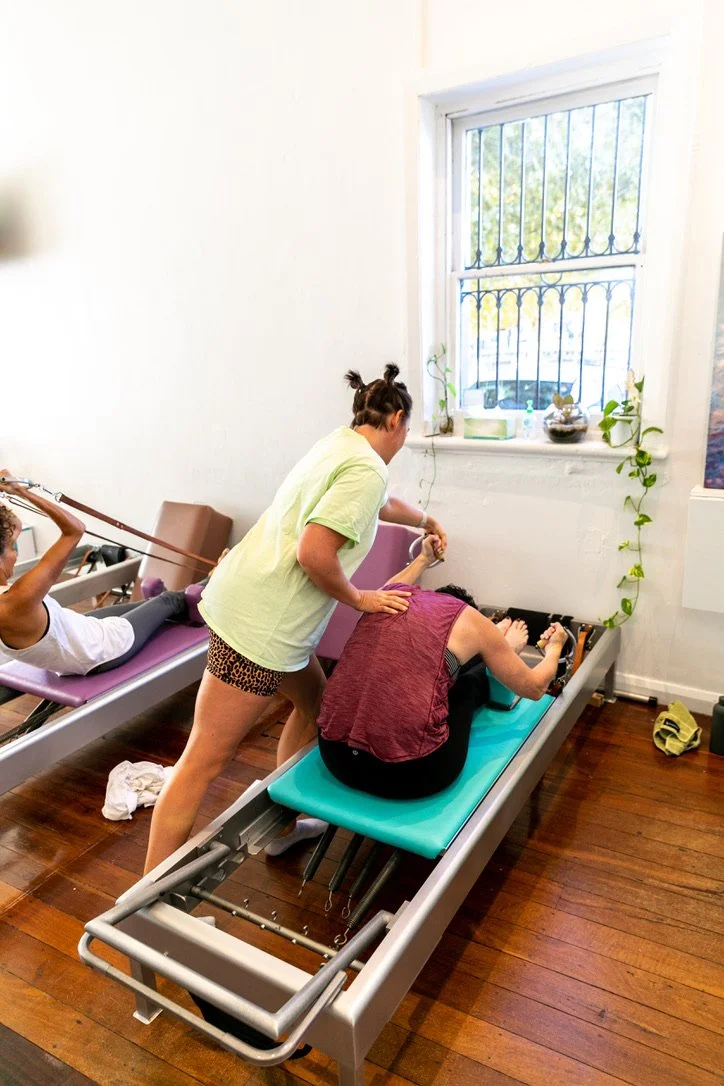 A woman on a Reformer is receiving assistance from a Pilates instructor.